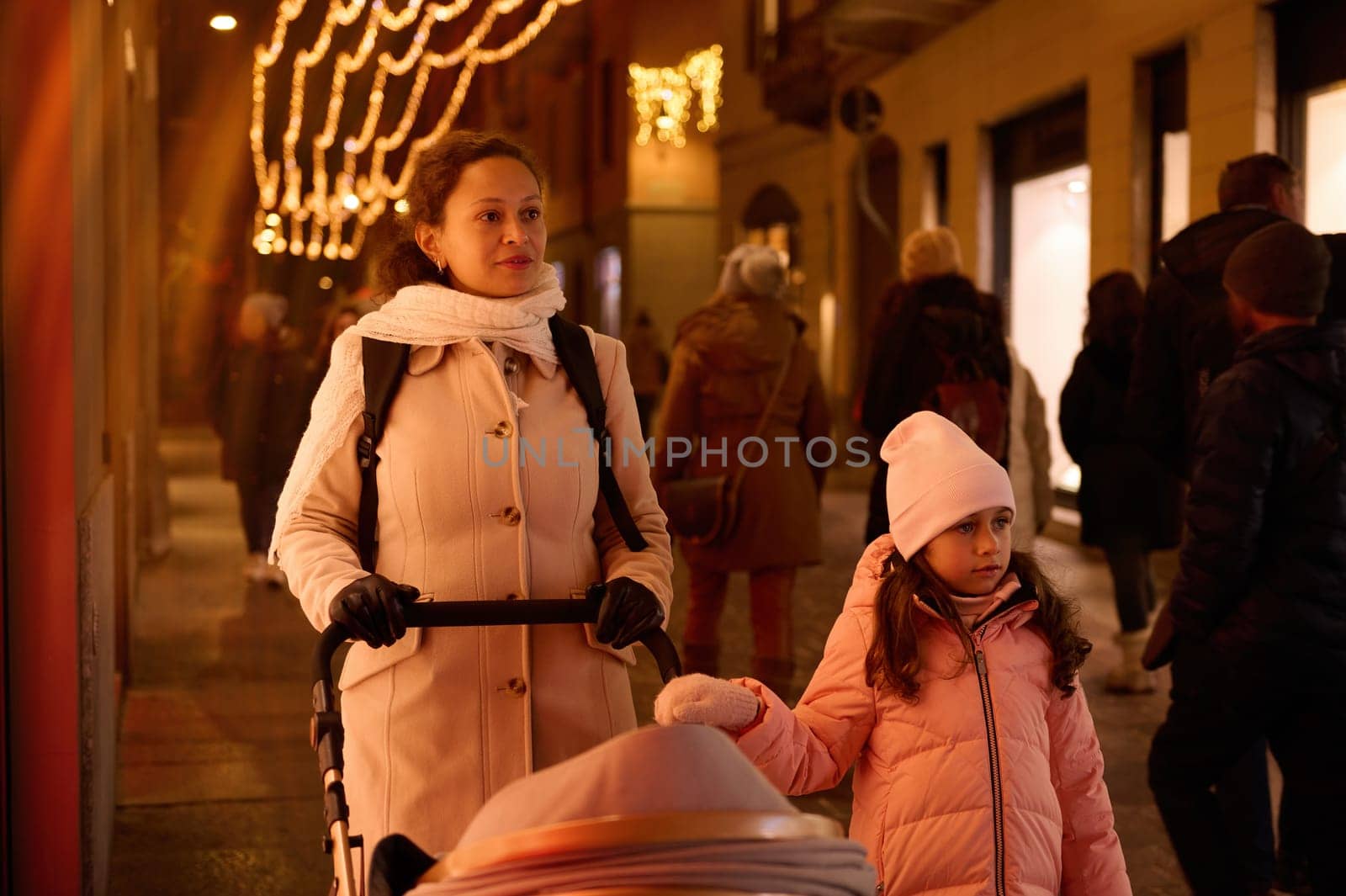Young mother pushing baby stroller, walking with her child daughter on the city street, enjoying festive atmosphere of Christmas fairground in the night time. People. Family. Holidays. Lifestyles