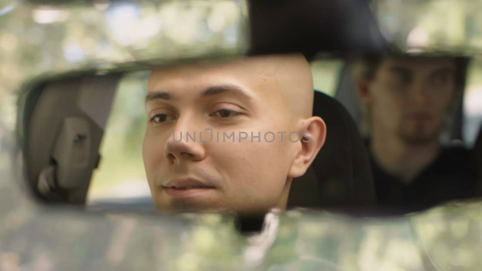Young man in rearview mirror of car. Stock. Man and friend are driving in car in summer. Bandit-looking men ride in car.