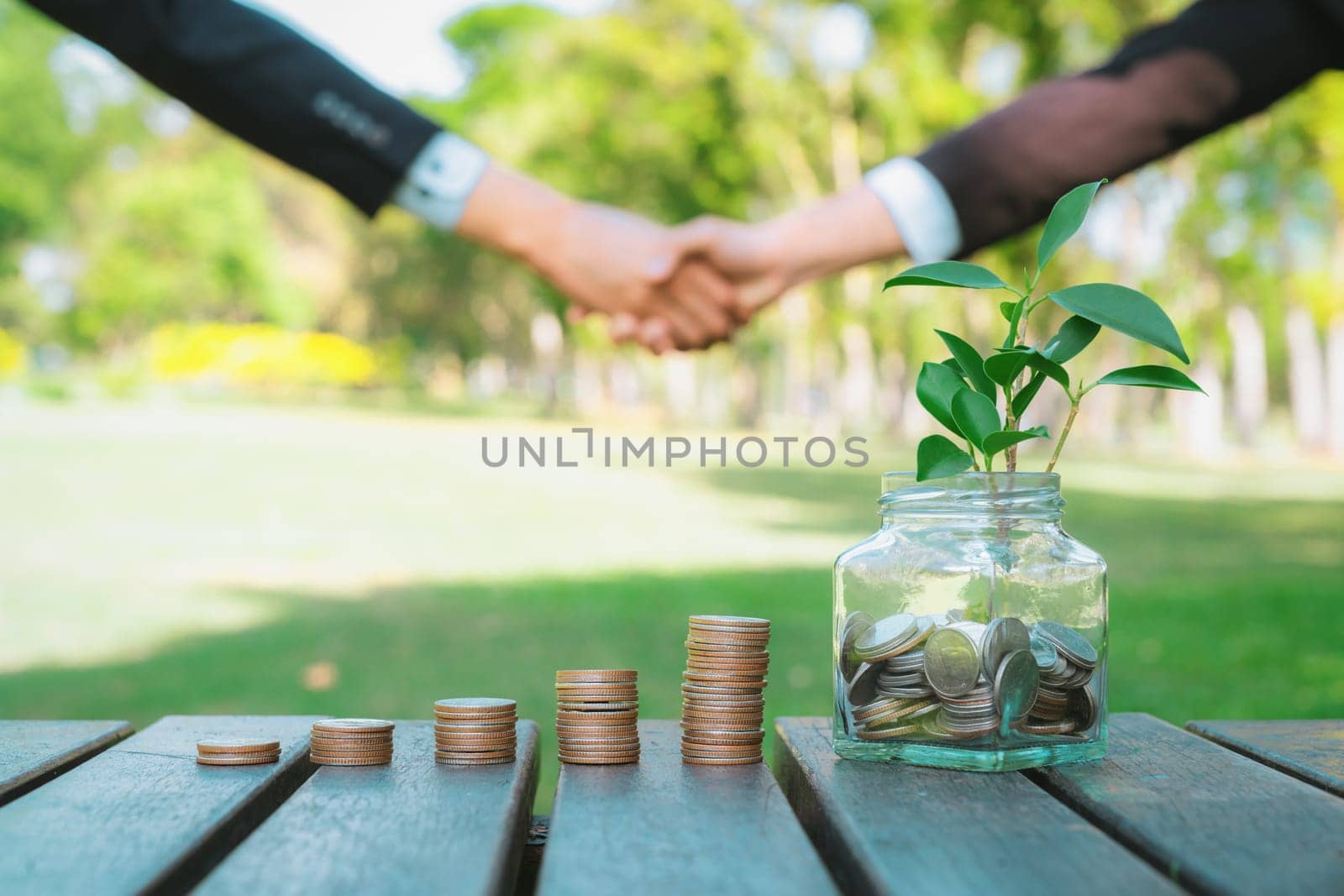 Sustainable money growth investment, glass jar filled with money saving and coin stack with businesspeople handshake in background as eco-friendly financial investment nurtured with nature. Gyre