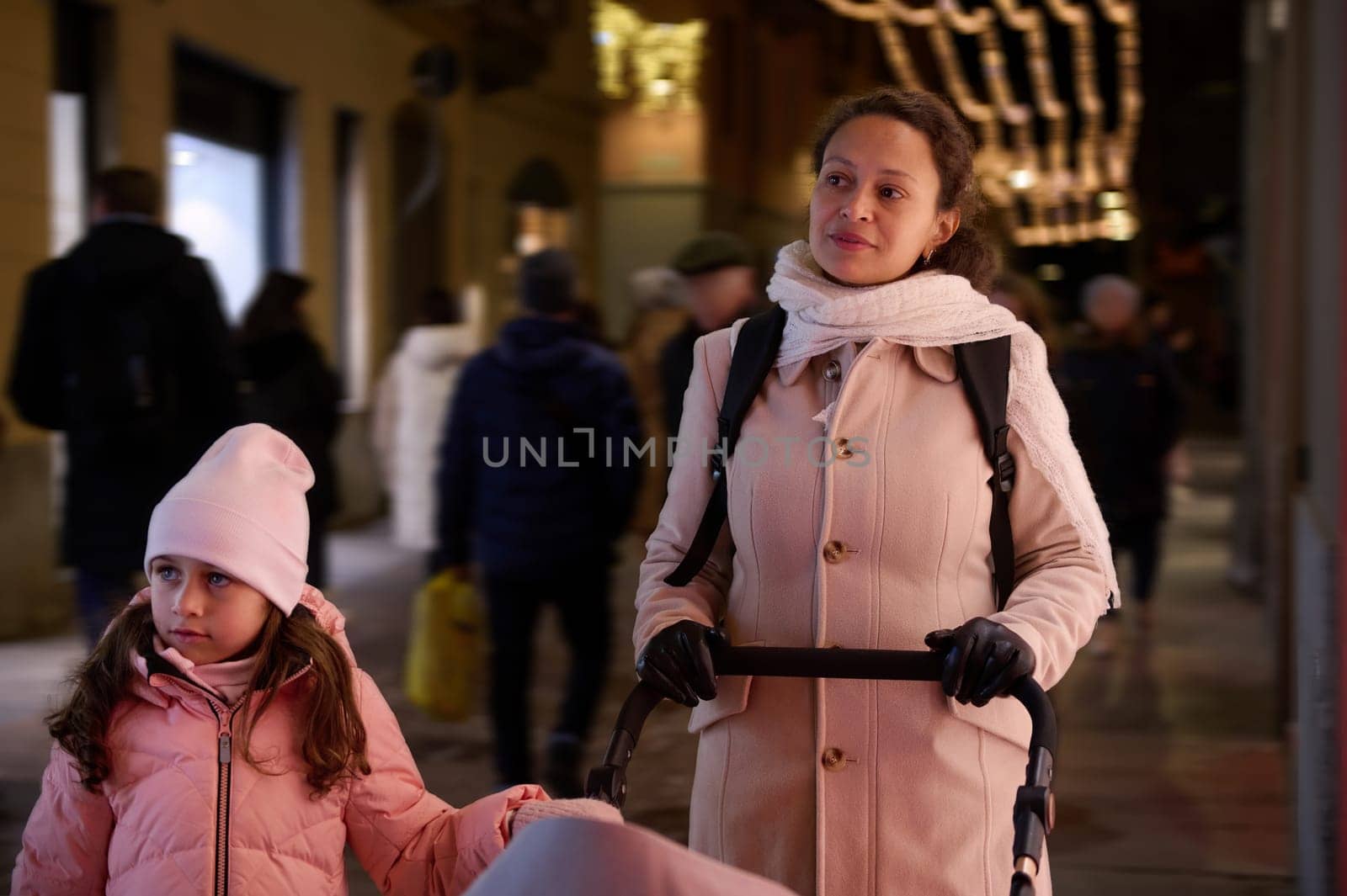 Mother with baby pram and her adorable little daughter strolling the street, enjoying festive atmosphere and Christmas funfair, visiting the traditional family market in December in the night time