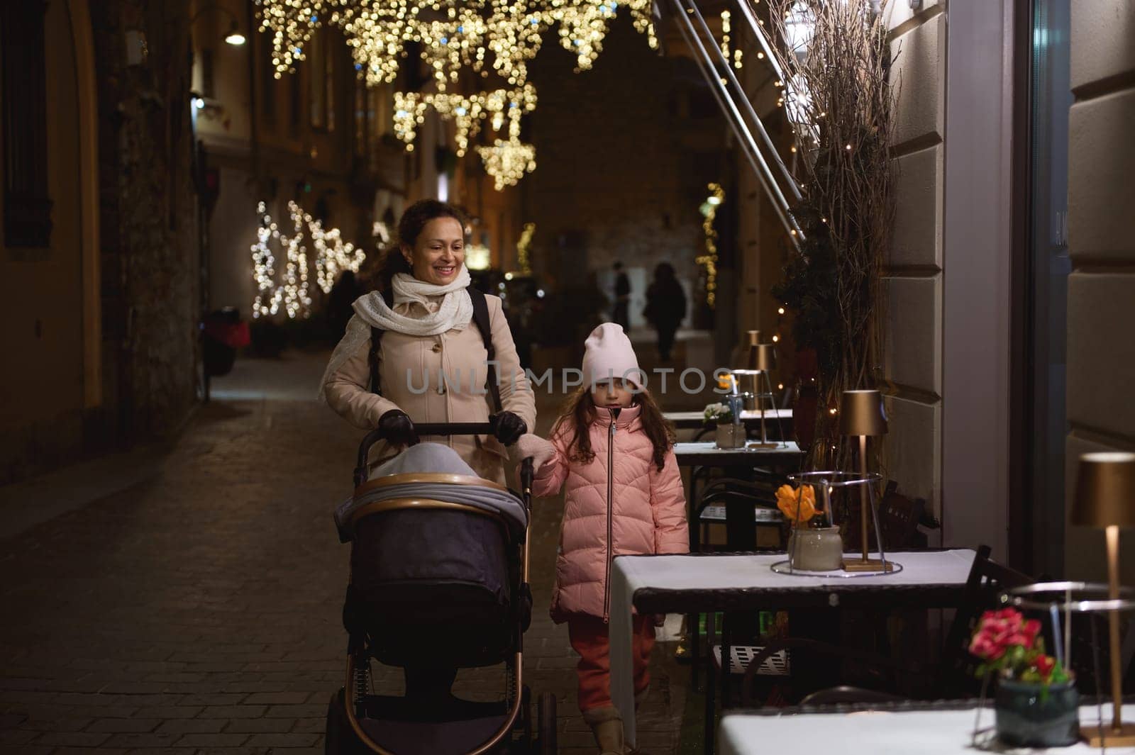 Authentic lifestyle portrait of a happy family of a mom holding the hand of her little daughter, pushing the baby pram, walking together on the street illuminated by festive lights in the night time