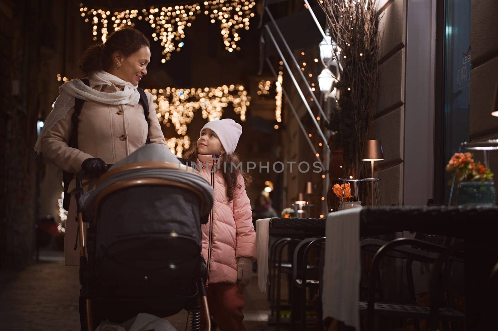 Happy young adult mother and daughter talking and smiling while strolling the street together, enjoying the walk in the night time during Christmas funfair. Family. Lifestyles. People. Winter holidays