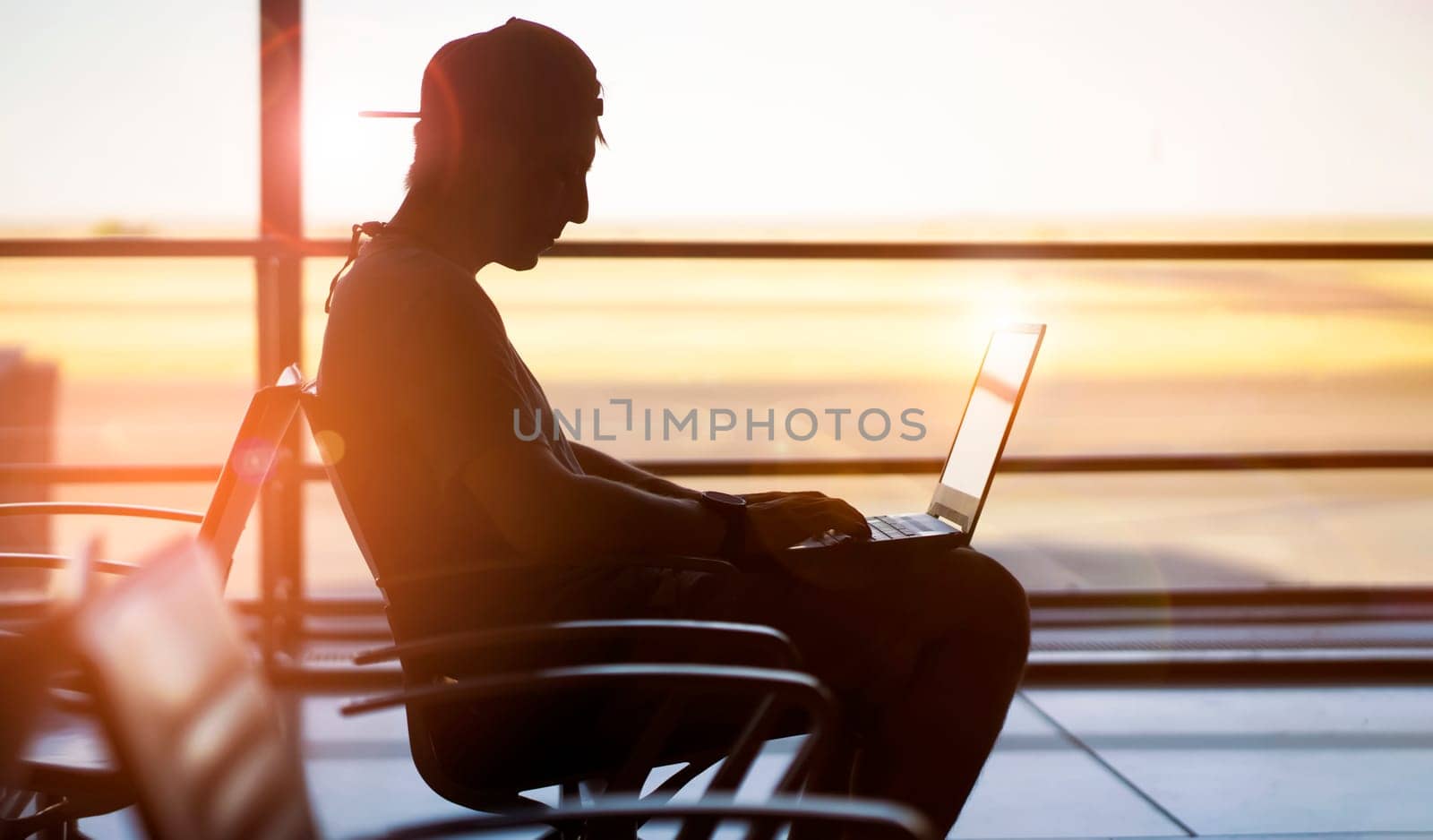 A young man is working on a laptop at the airport while waiting to board the plane. A man is engaged in business, buys tickets, studies and communicates via the Internet at sunset.