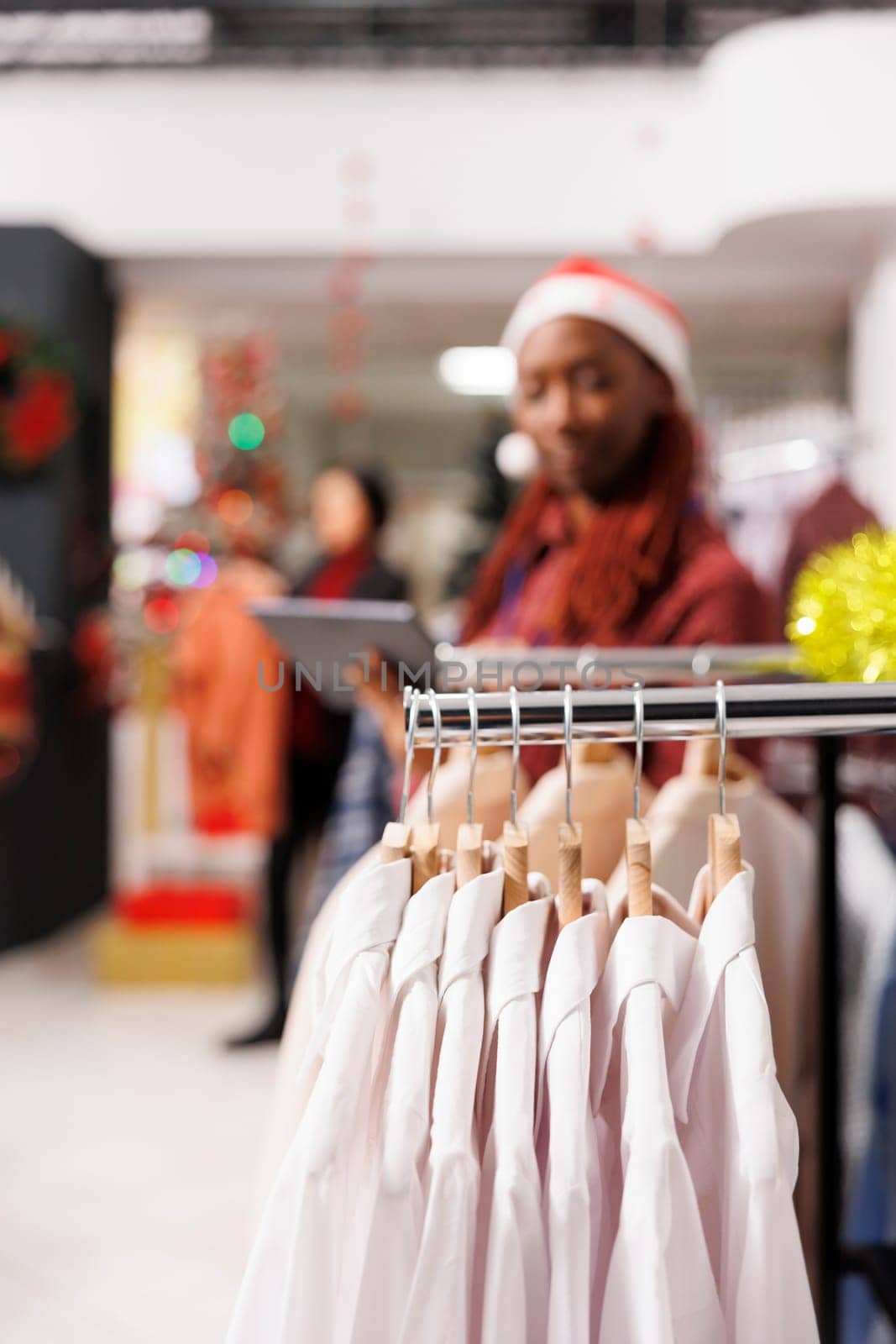 Selective focus of retail worker reviewing list of fashion items for stock inventory, working at mall during holiday season. African american employee managing logistics on tablet, xmas decor.