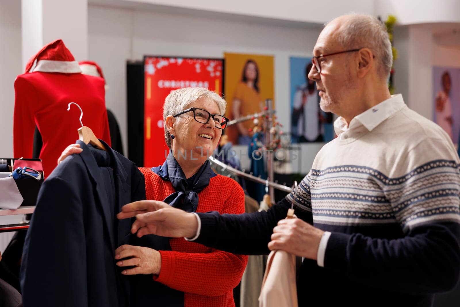 Customers checking suit jacket fabric in clothing store, reviewing blazers on racks and visiting mall to buy gifts on discount. Couple trying to choose fashion items on sale, holiday season.