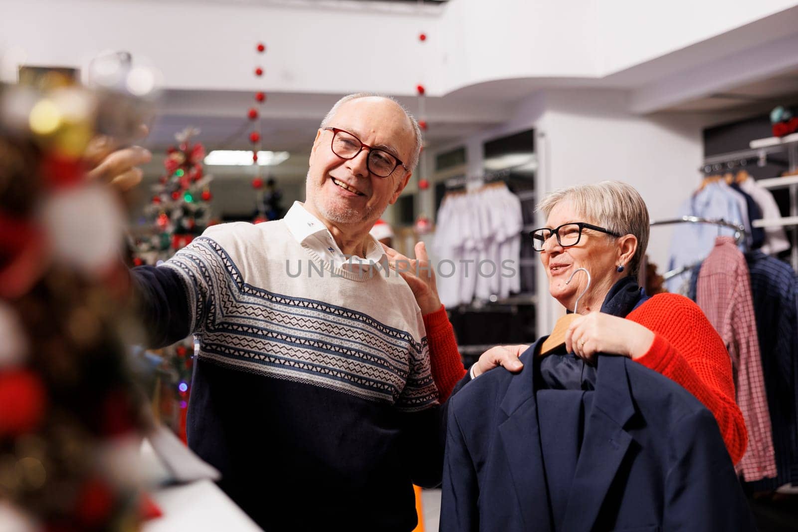 Senior couple searching clothing on videocall in retail store, trying to buy perfect fashion item measurement for christmas festivity. Two older people shopping for xmas gifts during holiday season.