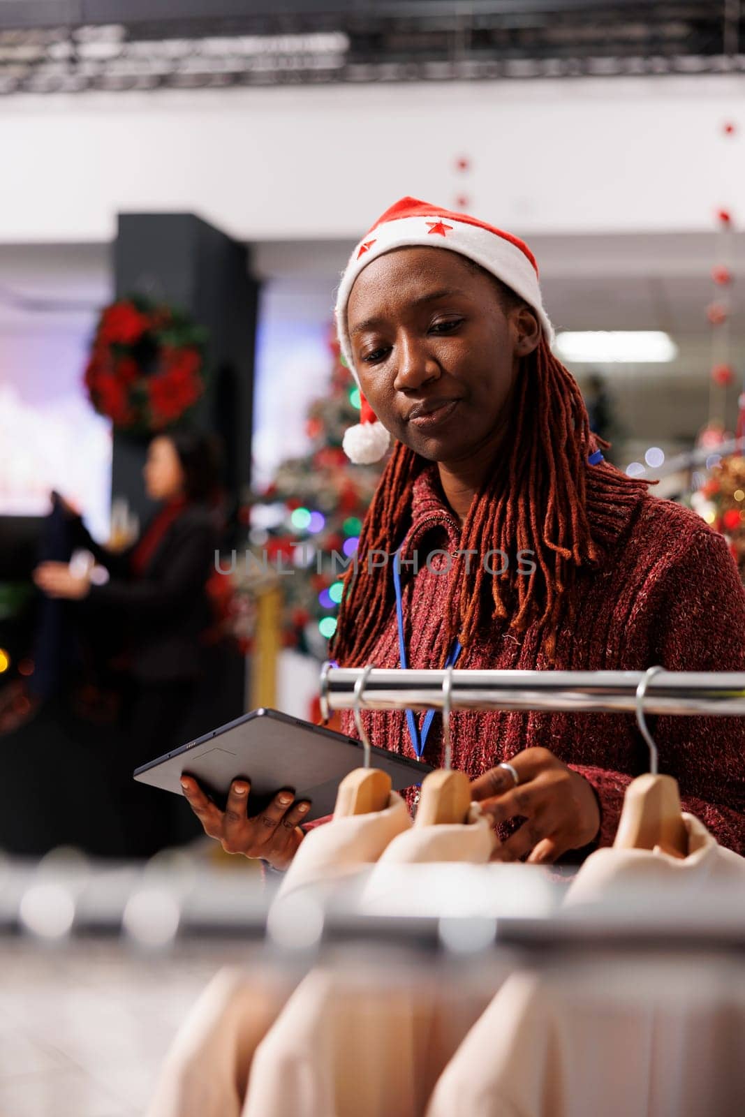 Sales manager checks inventory list with clothing items in retail store, working on stock during christmas holiday season. Woman with santa hat counting merchandise on racks, festive boutique.