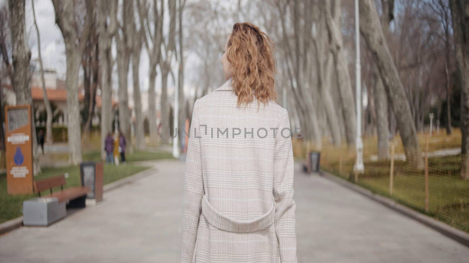 Rear view of a woman with curly hair walking in the city park in early autumn. Action. Young woman in coat enjoying her walk among bald trees