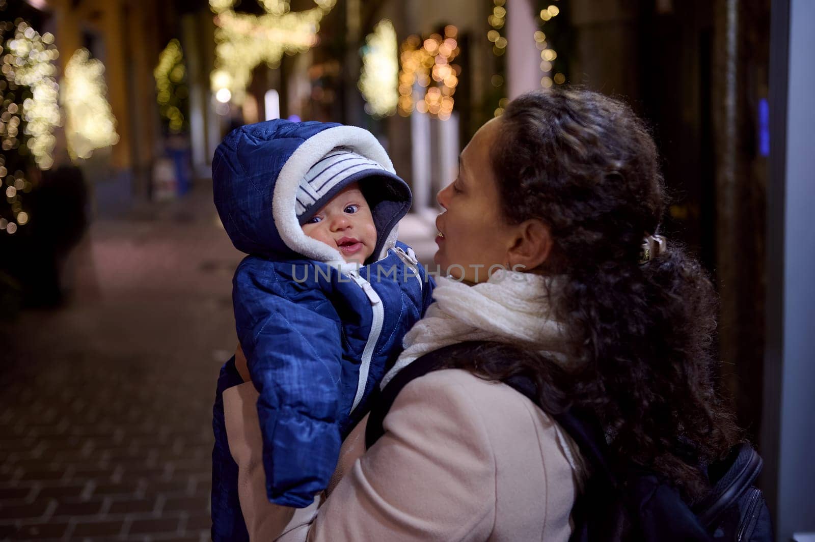 Beautiful young woman, mother holding her baby boy looking at camera, standing together in the street at night in elegant touristic city Como of Italy, decorated with beautiful garlands for Christmas