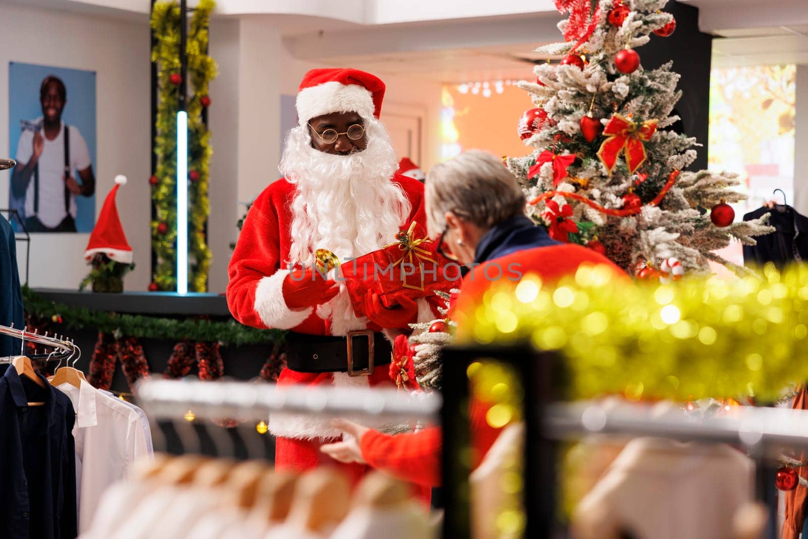 Santa Claus man in suit holding raffle at shopping center, inviting elderly person to participate at contest to win free clothing item during christmas holiday season. Festive decorated retail store.
