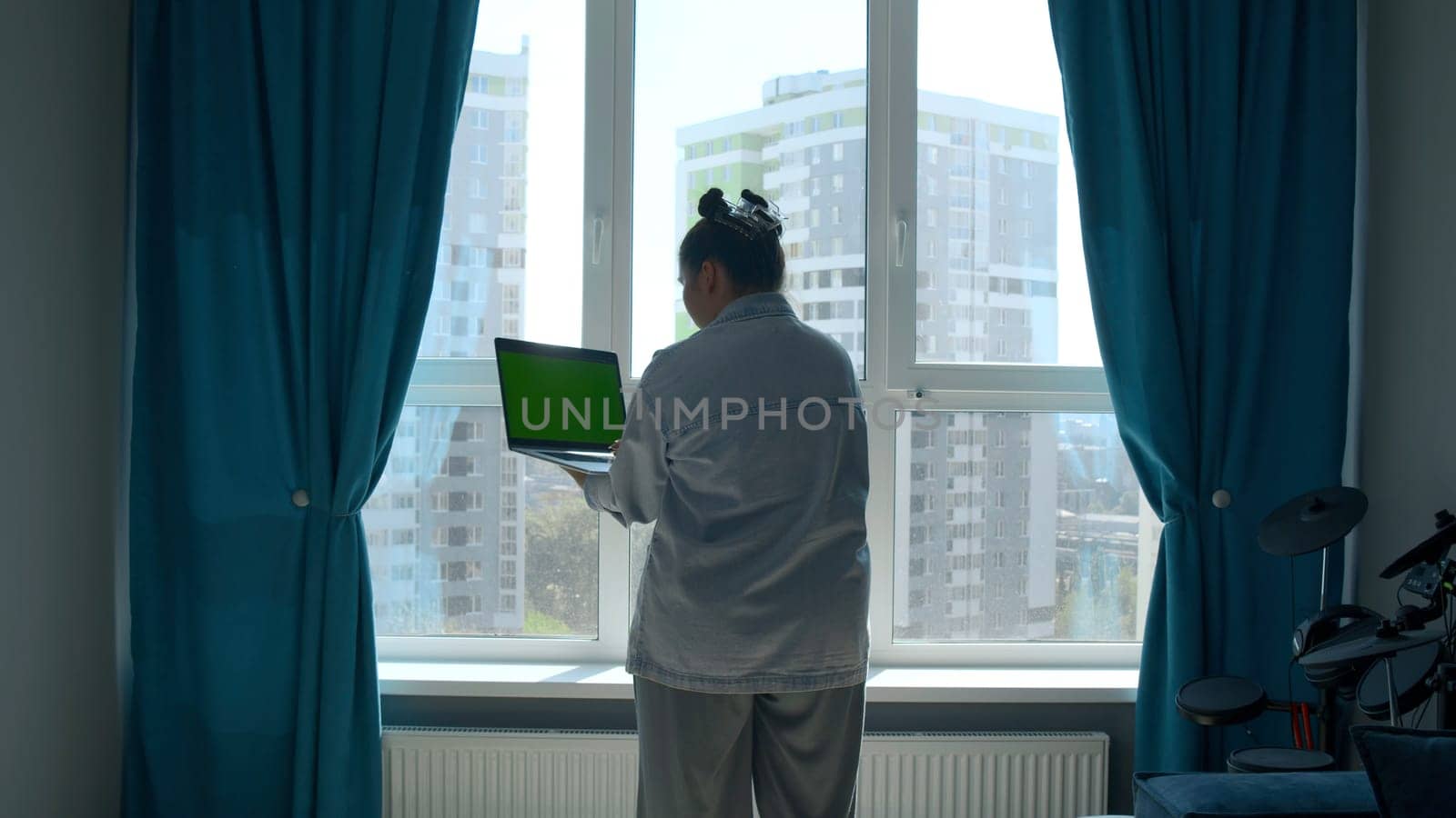 Rear view of a woman with funny hairstyle standing by the window with a laptop in her hands. Media. Computer with green chroma key screen