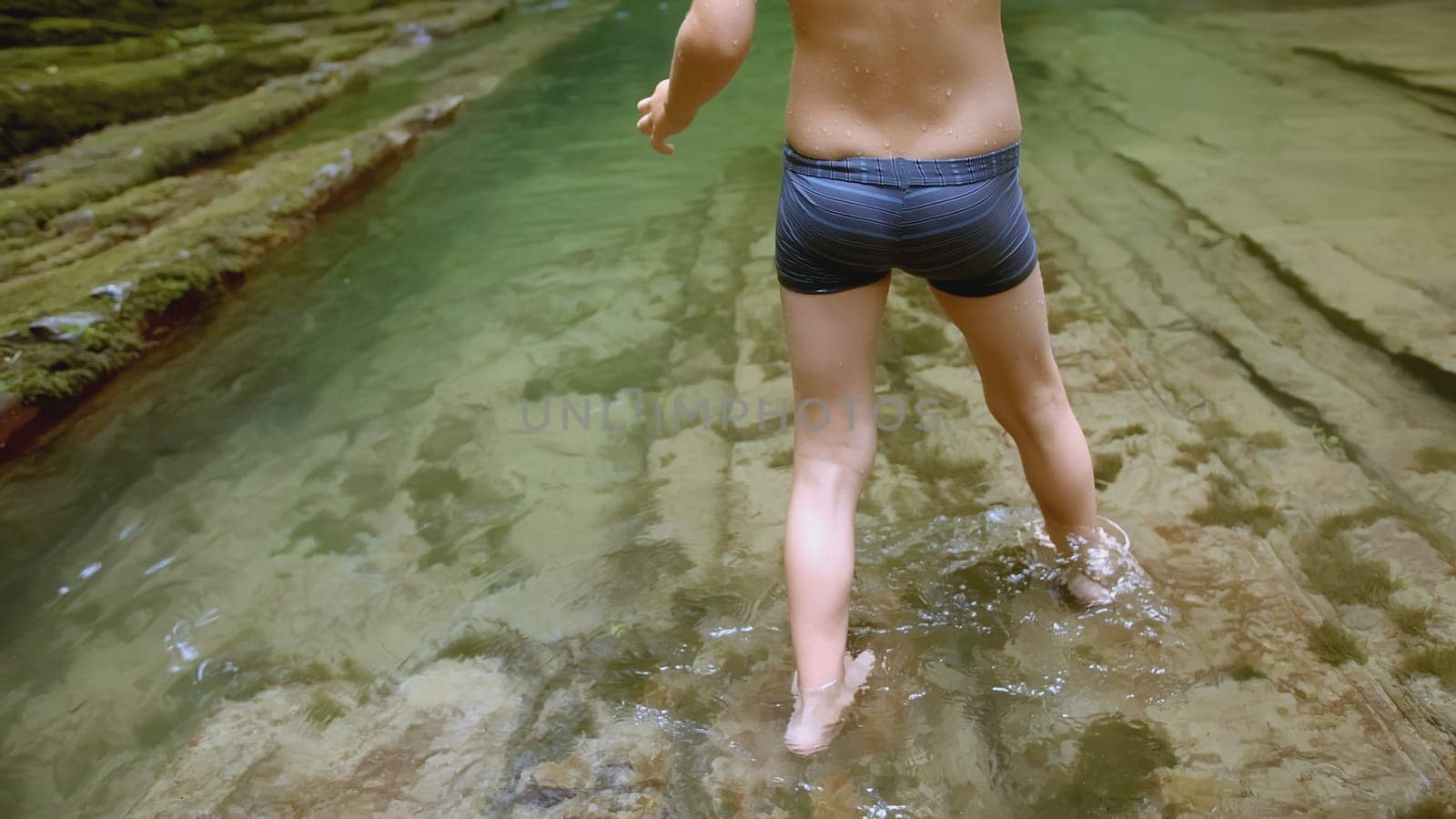 Boy walks in water in forest. Creative. Boy in swimming trunks walks on water in summer forest. Boy's feet walk on water in wild forest in summer. Summer hike in wild woodland by Mediawhalestock