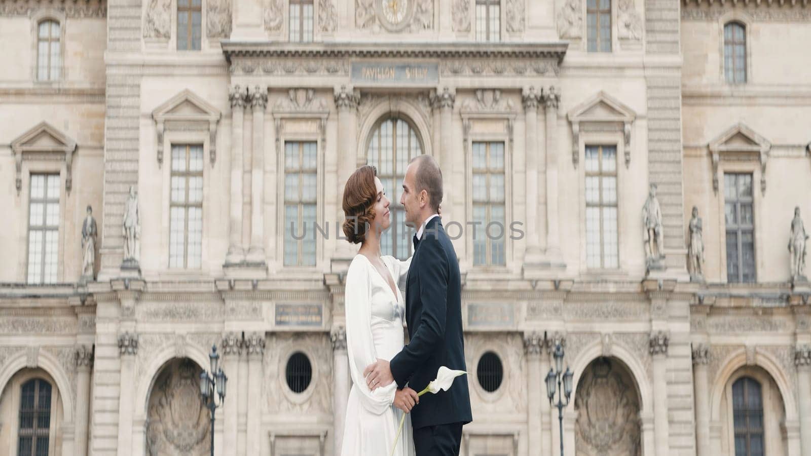 Adult couple in elegant outfits hugging. Action. Luxurious couple embraces in love on background of palace. Couple in love in elegant outfits on old square by Mediawhalestock