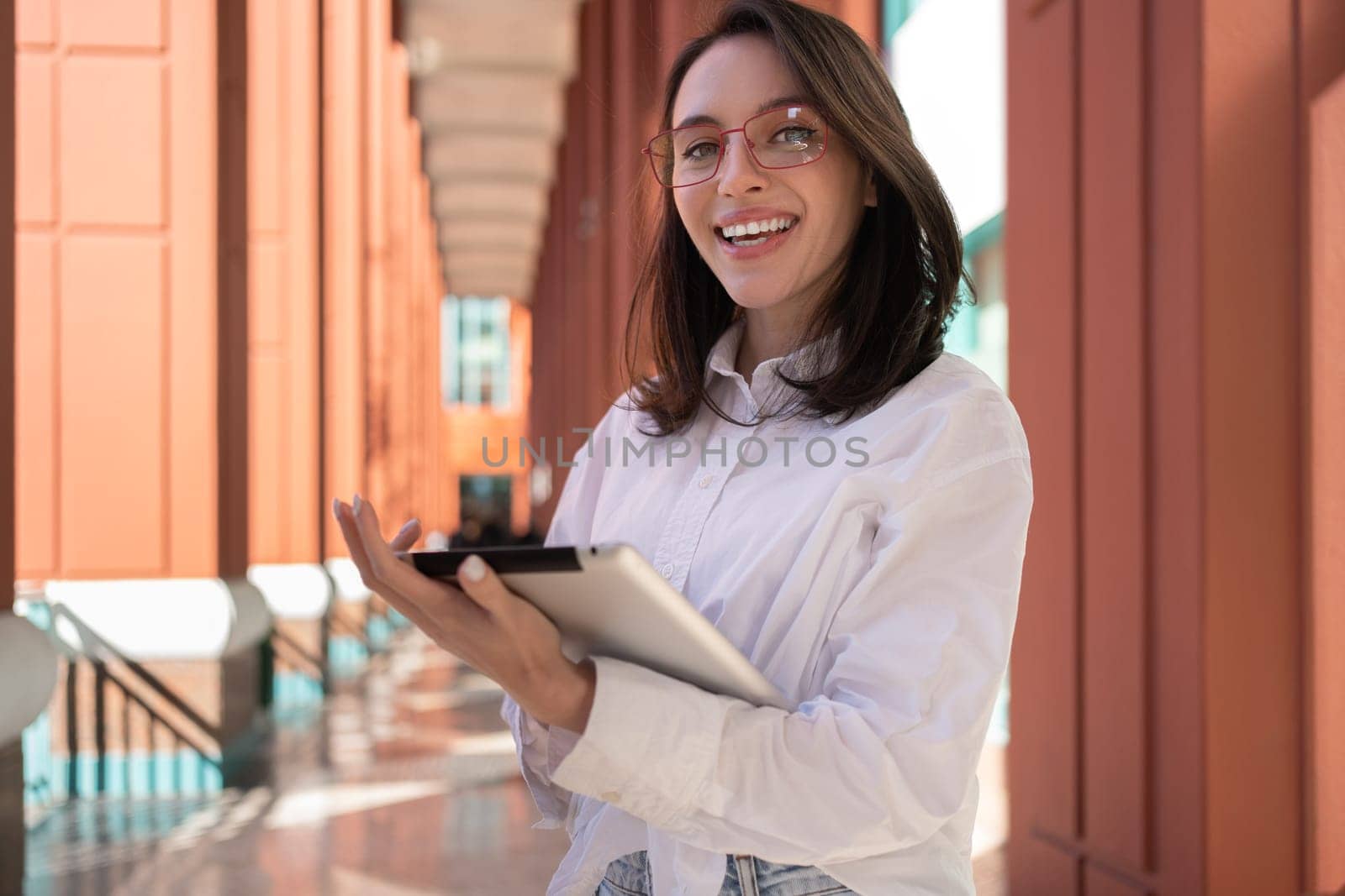 Woman standing with tablet wear glasses smiling at camera near office. Businesswoman dressed in smart casual clothes holding digital tablet and smiles at camera on street near building. Blur background