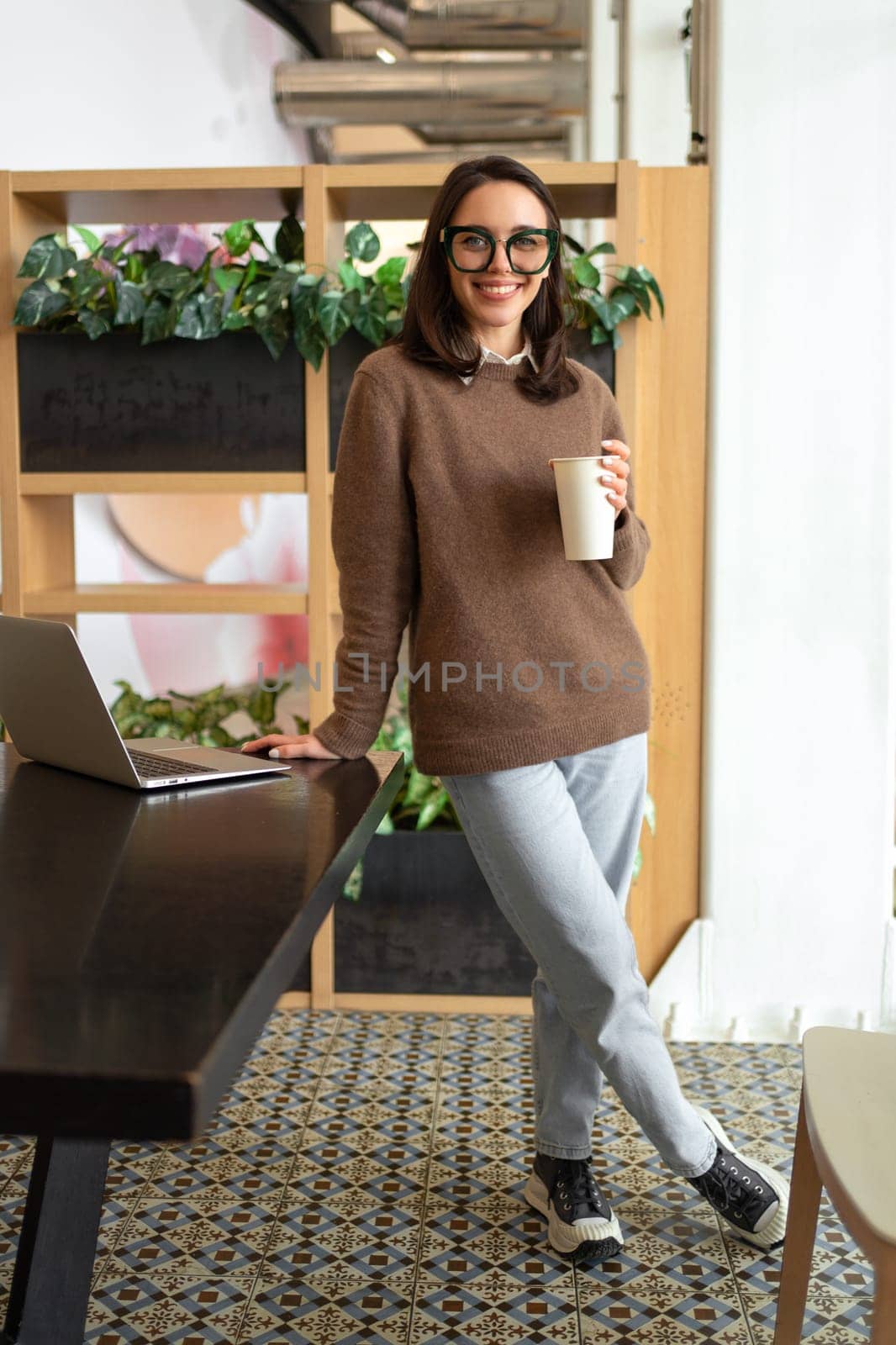 Woman in glasses poses smiling standing at workplace with cup coffee. Woman dressed in smart-casual is standing leaning on table and smiling. Female stands by work laptop happy to be relaxing