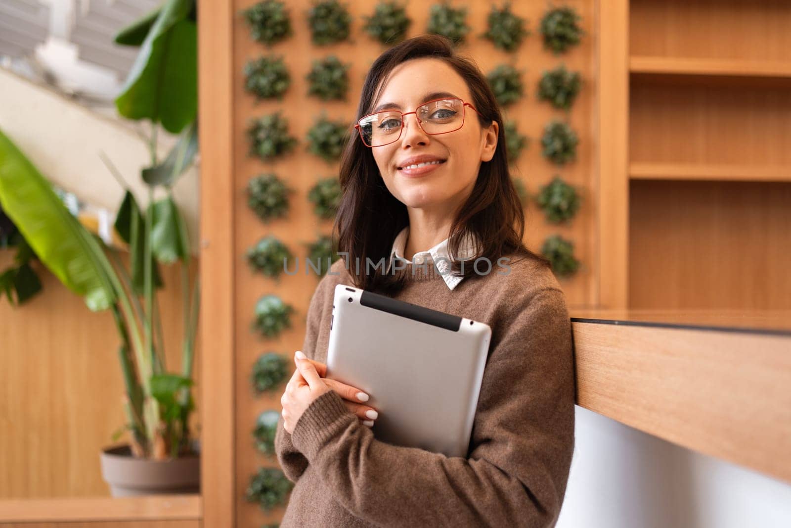 Confident woman standing in eyeglasses and tablet on hands. Confident business woman standing on hall looking at camera holding digital tablet at blur background. Employee poses for photo