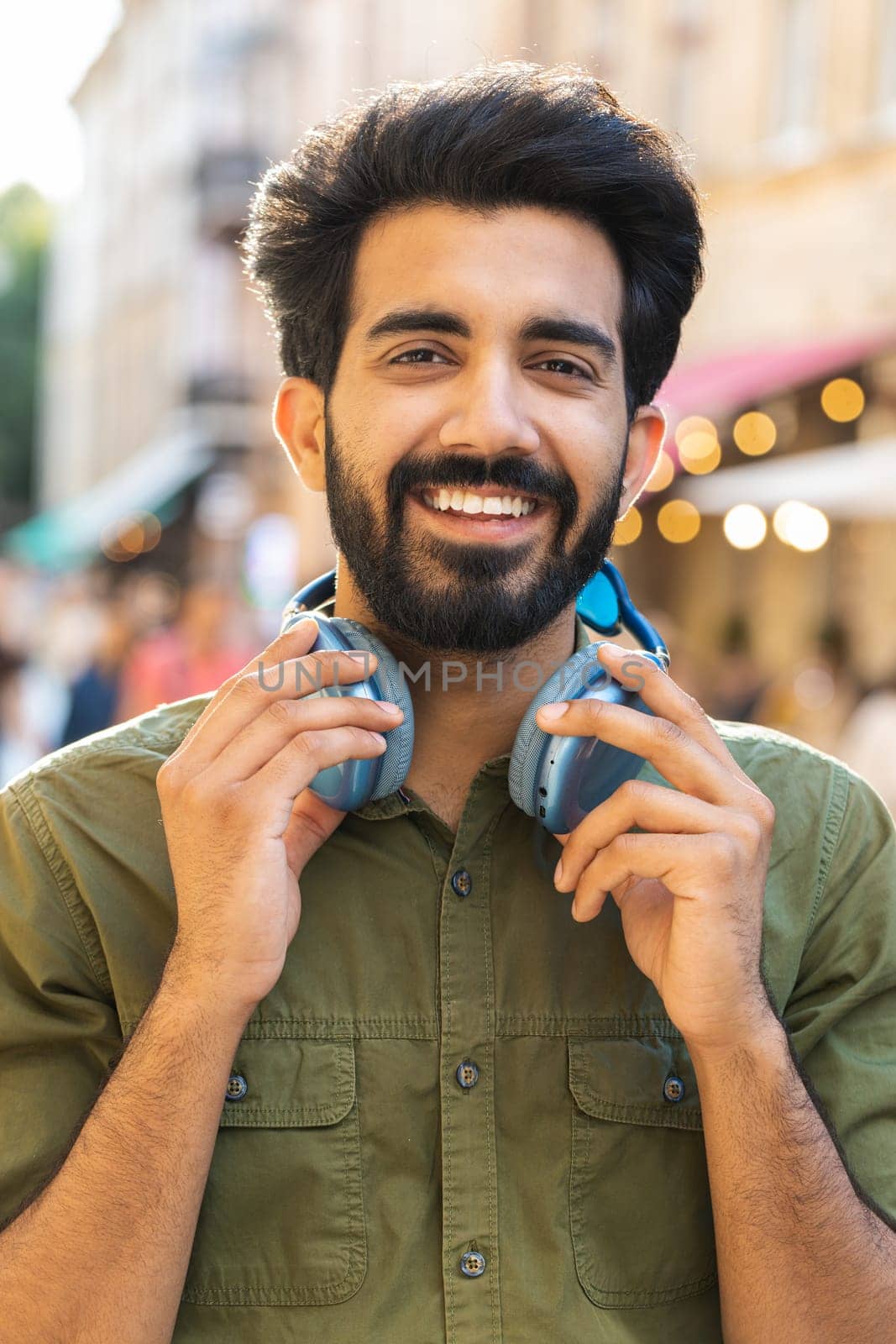 Joyful relaxed overjoyed indian man choosing, listening favorite energetic disco rock n roll music dancing outdoors. Guy taking off wireless headphones looking at camera in urban city street. Vertical