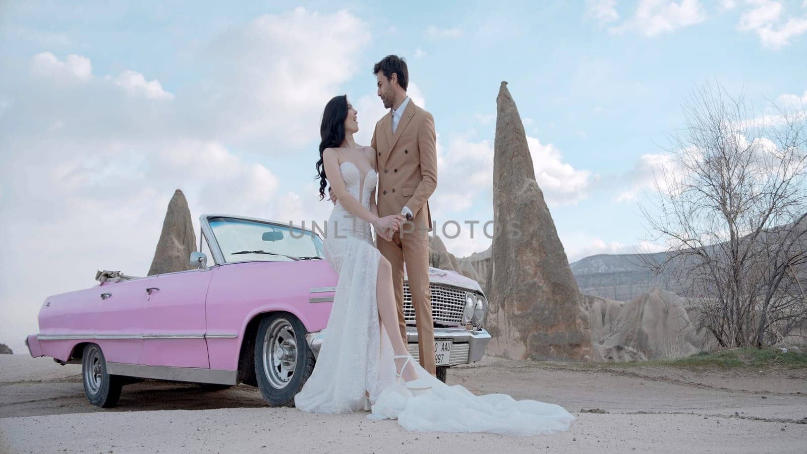 Happy bride in long white dress and groom in brown suit, newlywed wedding couple standing by convertible pink car. Action. Cabriolet with stone formations on the background