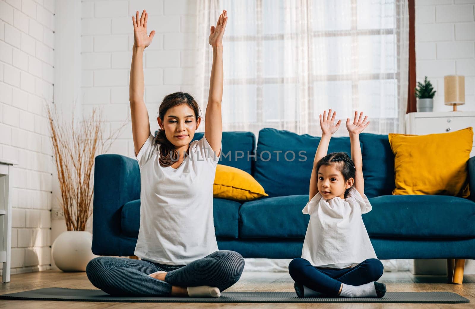 A beautiful young woman and her charming daughter share smiles during their family yoga session at home focusing on stretch and balance creating a harmonious and joyful moment.