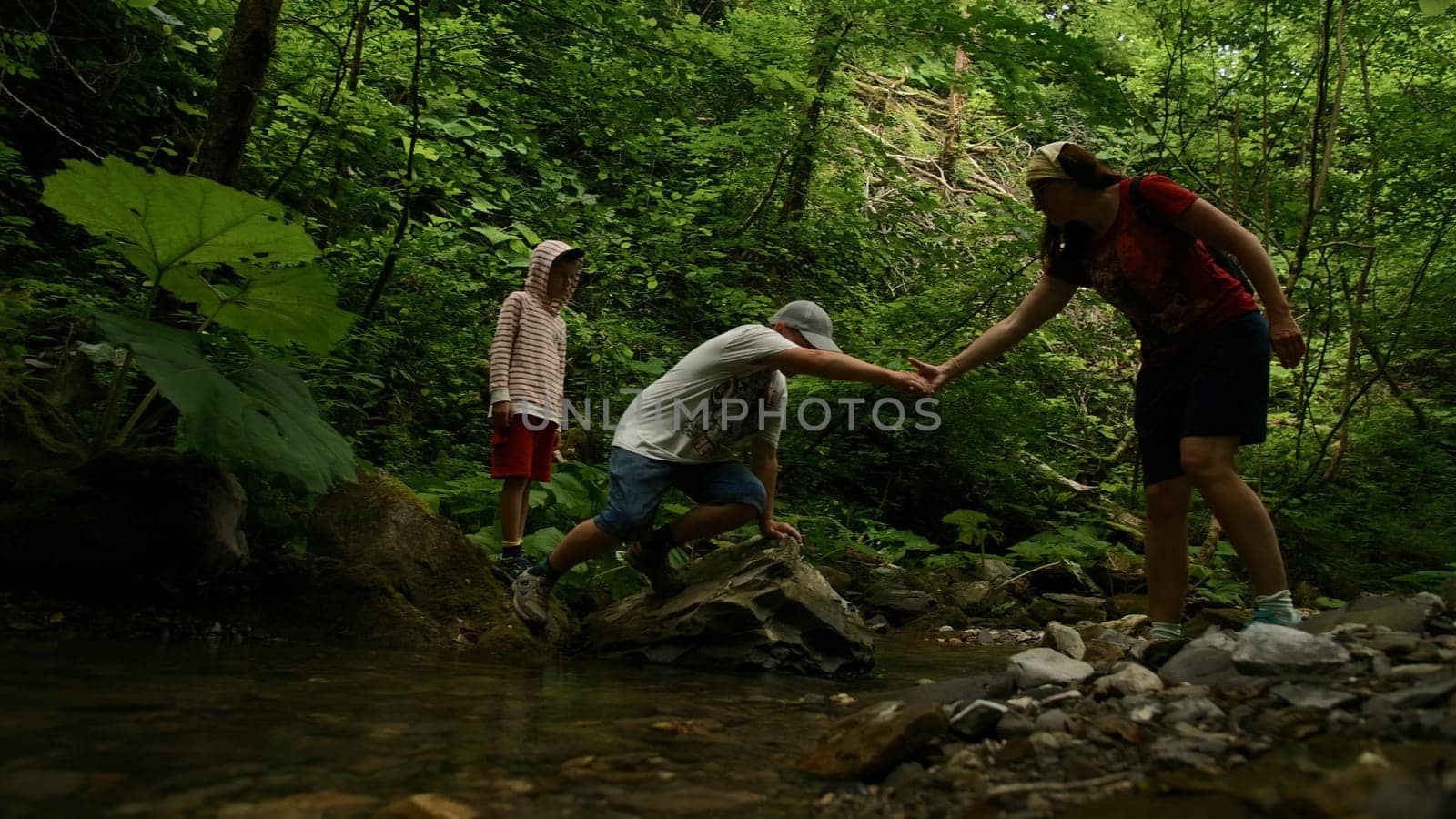 Family crossing narrow stream with stones. Creative. Hikers enjoying beautiful landscape. by Mediawhalestock