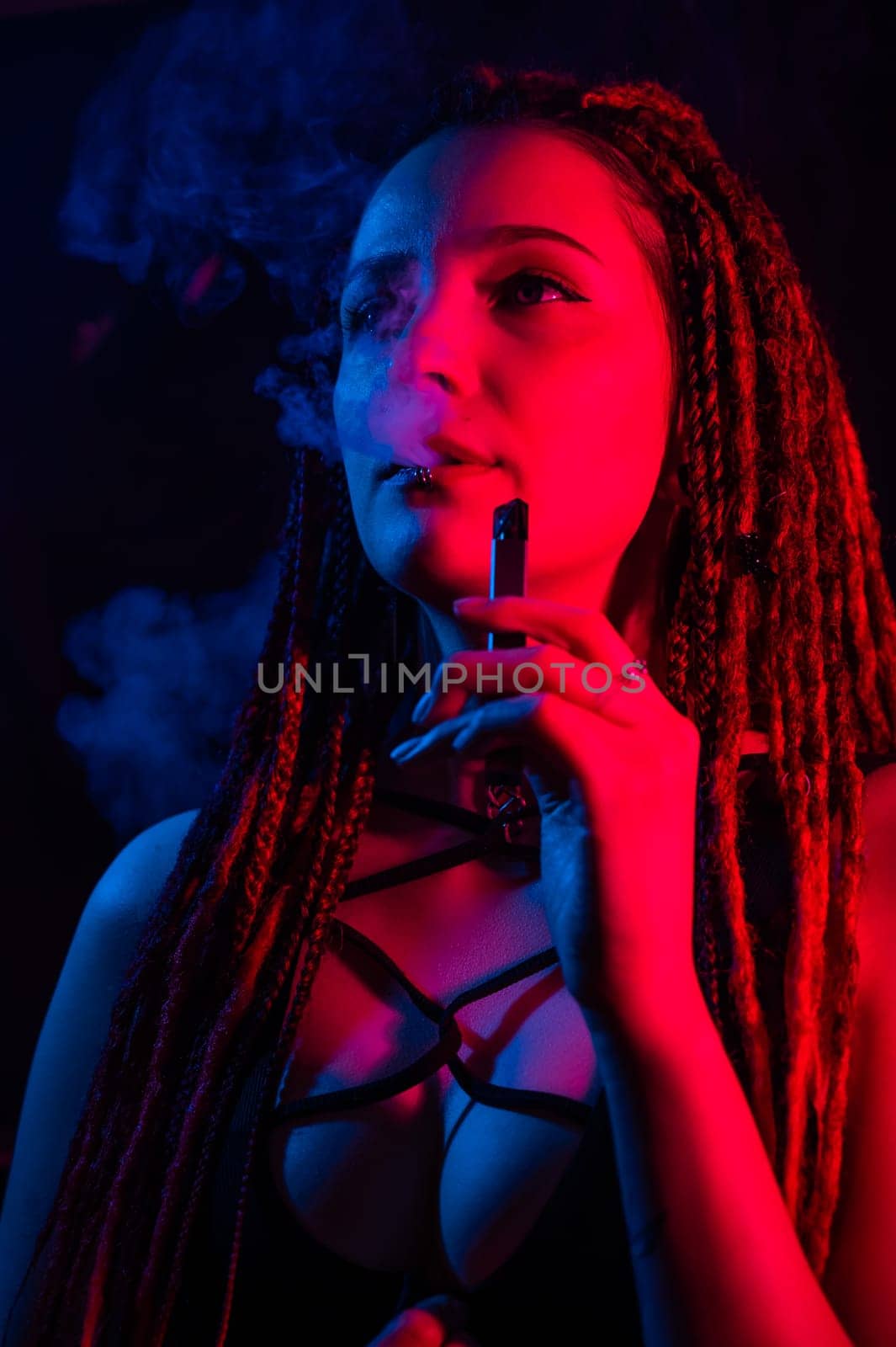 Caucasian girl with dreadlocks smokes a vape in red blue light