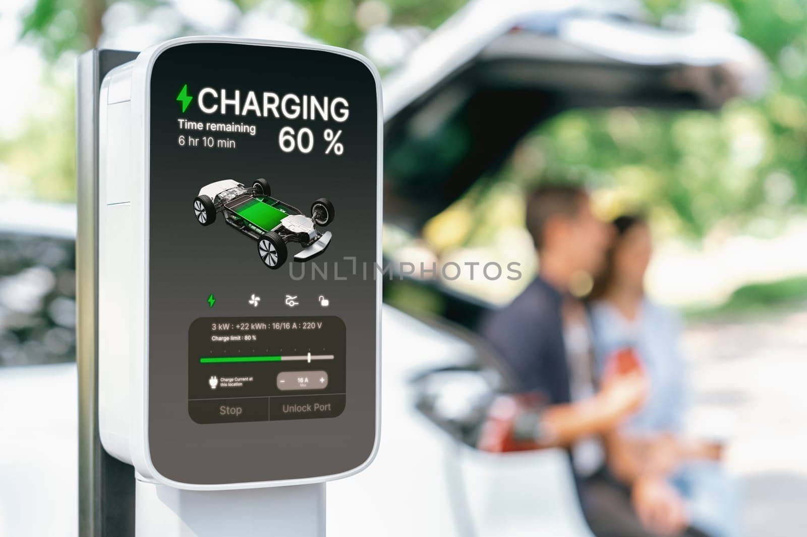 Focused EV charging station monitor display battery status for electric car on blurred background of couple sitting on car's trunk while recharging battery during natural travel and holiday. Exalt