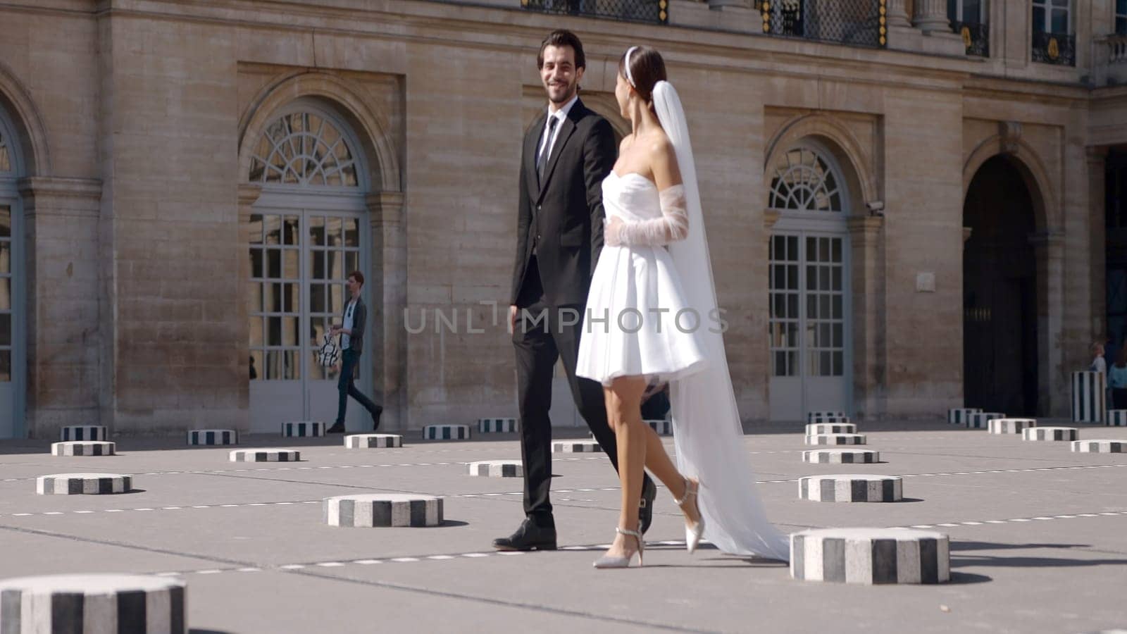 Beautiful newlyweds walking in city on sunny day. Action. Stylish wedding dresses for newlyweds walking on square. Happy newlyweds walk hand in hand in city square.