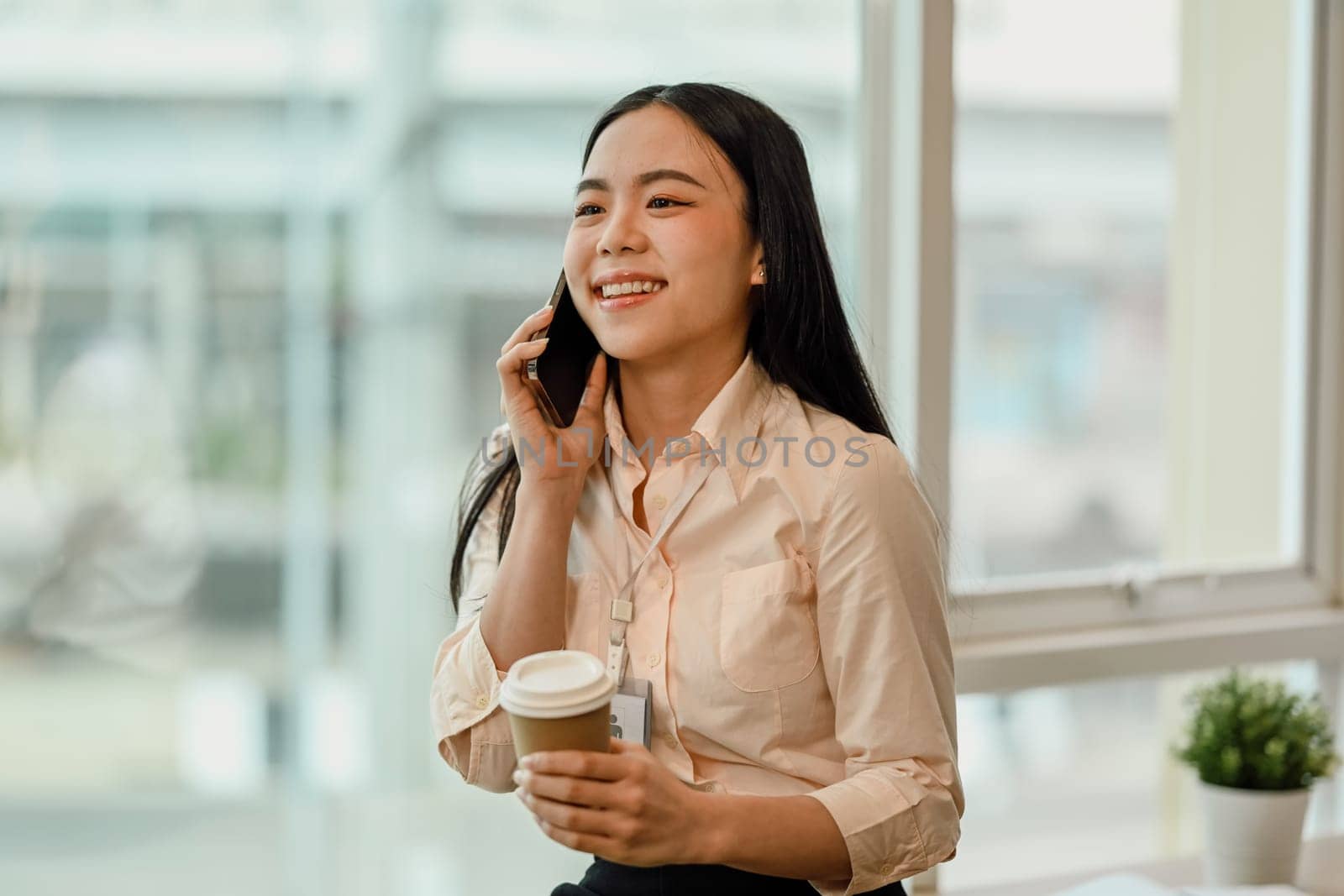 Cheerful Asian female employee talking on mobile phone near office window. by prathanchorruangsak