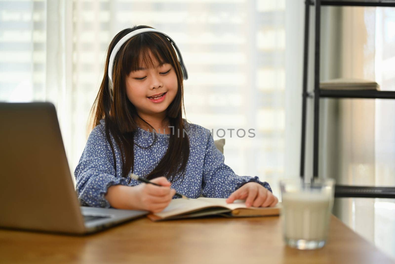 Smiling Asian schoolgirl in headphone studying online on laptop, doing homework at dinning table