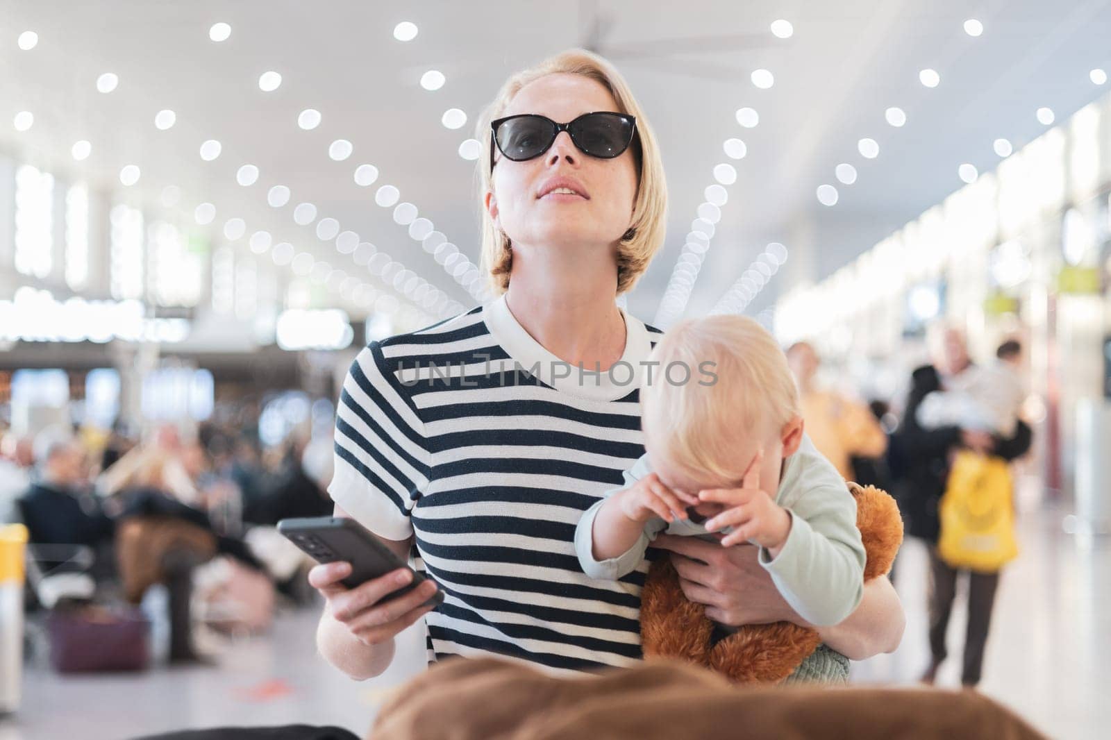 Mother traveling with child, holding his infant baby boy at airport terminal, checking flight schedule, waiting to board a plane. Travel with kids concept