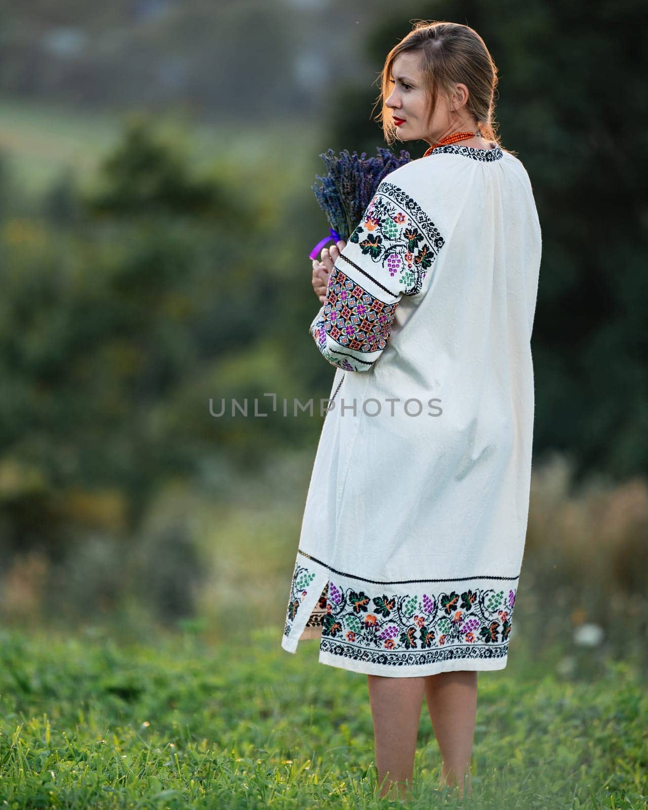 A young girl wearing a long embroidered shirt holds a bouquet of lavender in her hands. by Niko_Cingaryuk