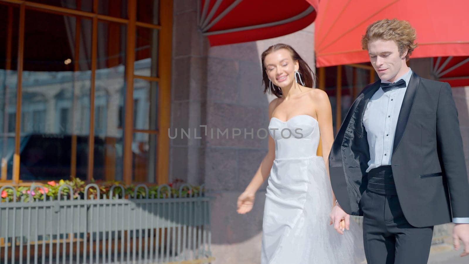 Wedding photo shoot on the street.Action.A bright and spectacular couple with a bride in a white dress with a neckline and long earrings and a man in a black suit on the street next to buildings. by Mediawhalestock