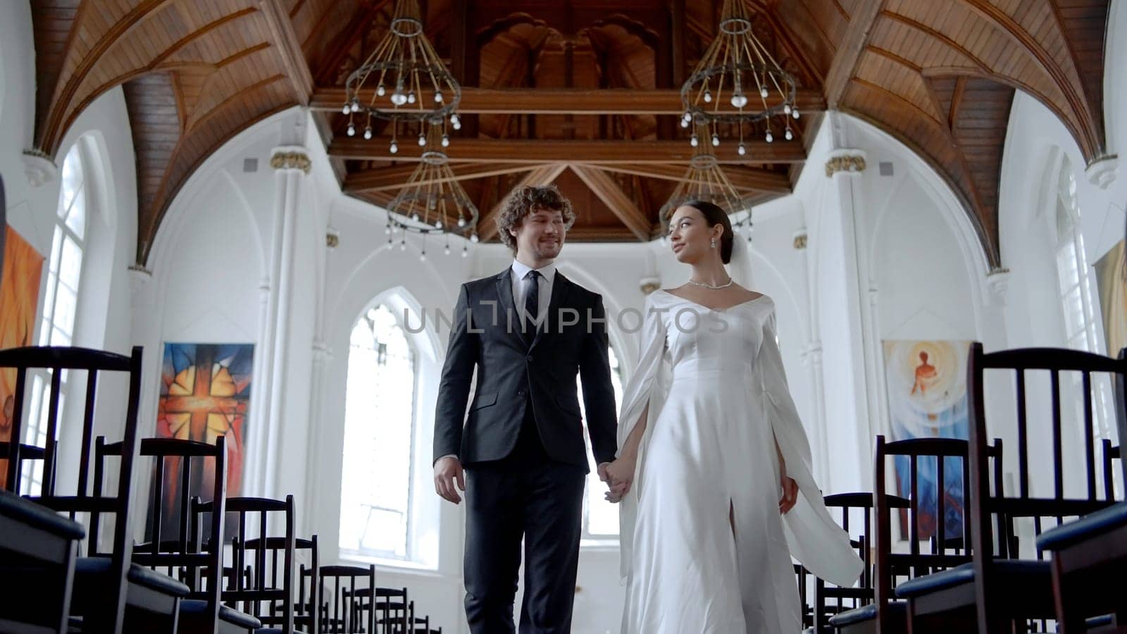 A couple of newlyweds walking quickly through the temple. Action.A beautiful bride in a white tight dress and a veil with a man in a suit with a beard are running with a quick gait through the church hall. by Mediawhalestock