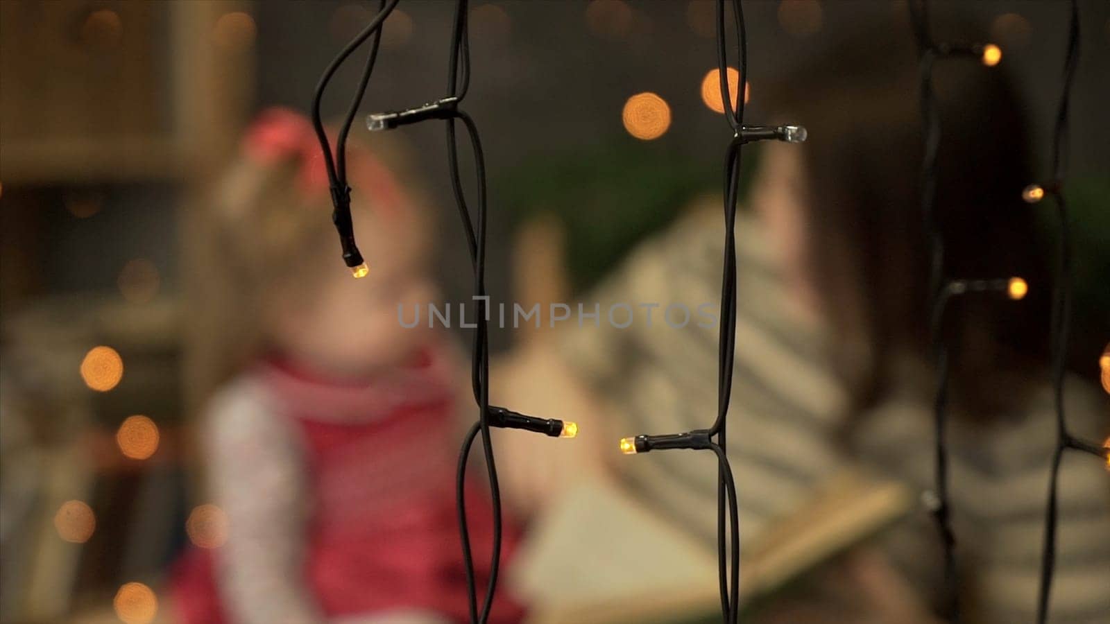 Young mother with little daughter reading book at home. Mom and little daughter playing on background of garlands. Blur by Mediawhalestock