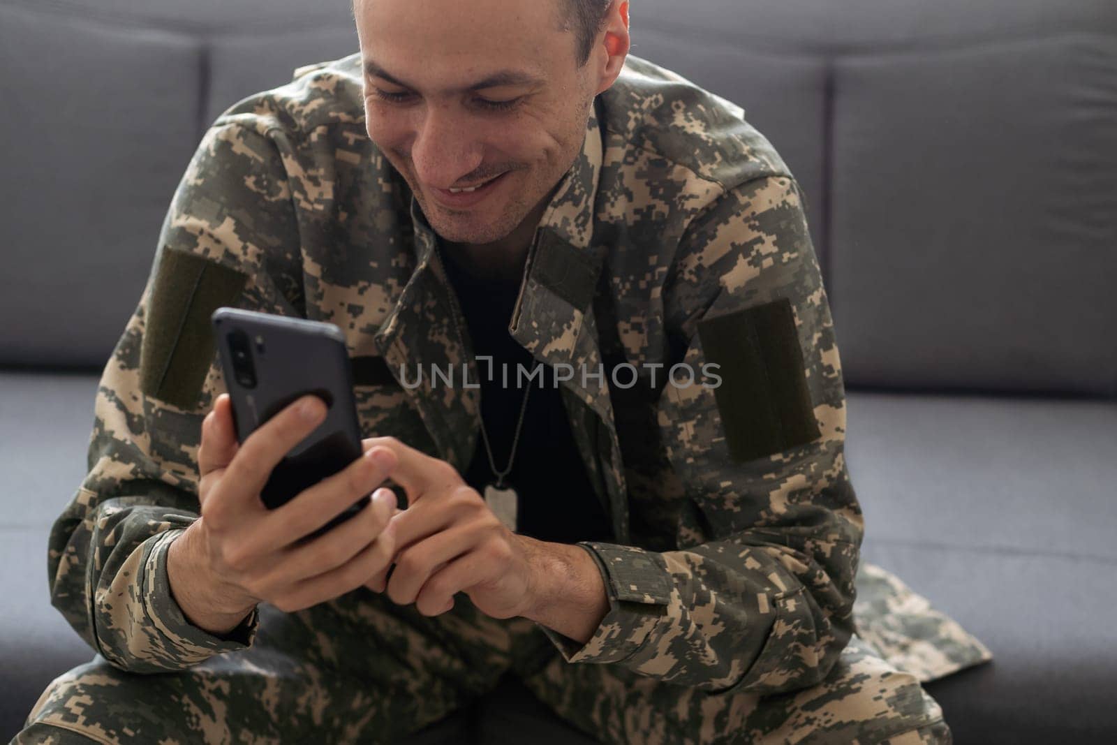 Portrait of smiling positive military man wearing camouflage uniform sitting on sofa, soldier talking on video call, saying hello or bye, looking at device screen with positive expression. High quality photo