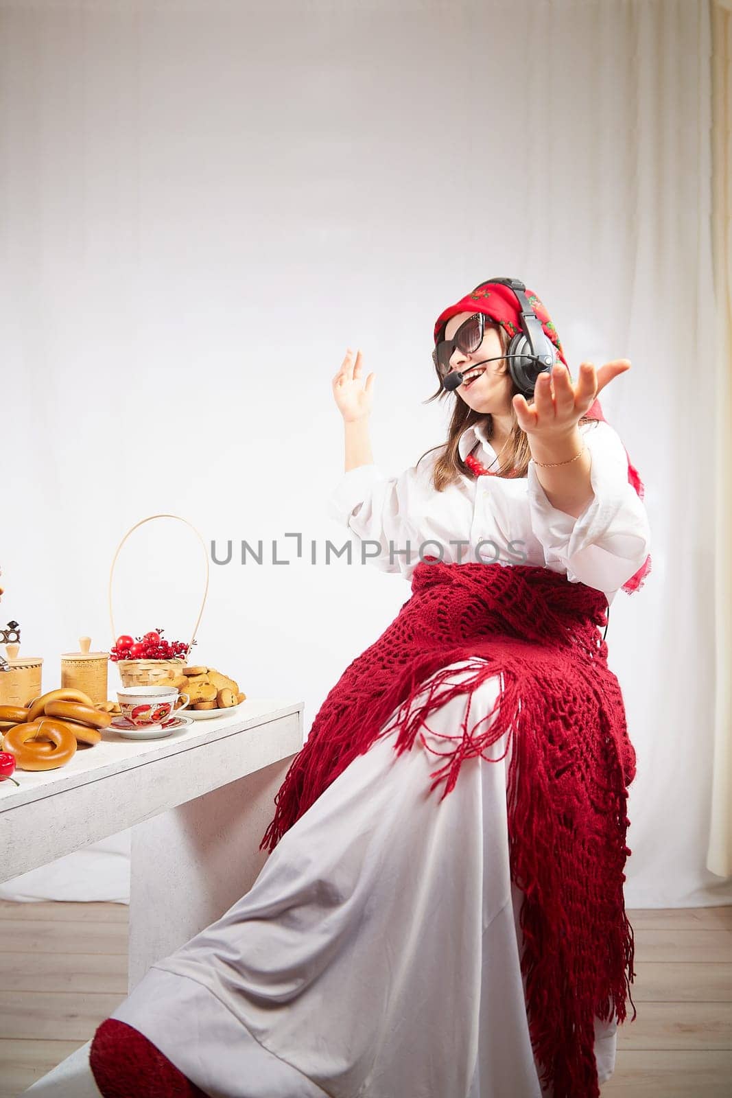 Girl in red scarf and large headphones with microphone having fun at table with delicious food for Orthodox holiday Maslenitsa or Easter. Funny Woman operator, blogger, Freelancer singing and dancing by keleny