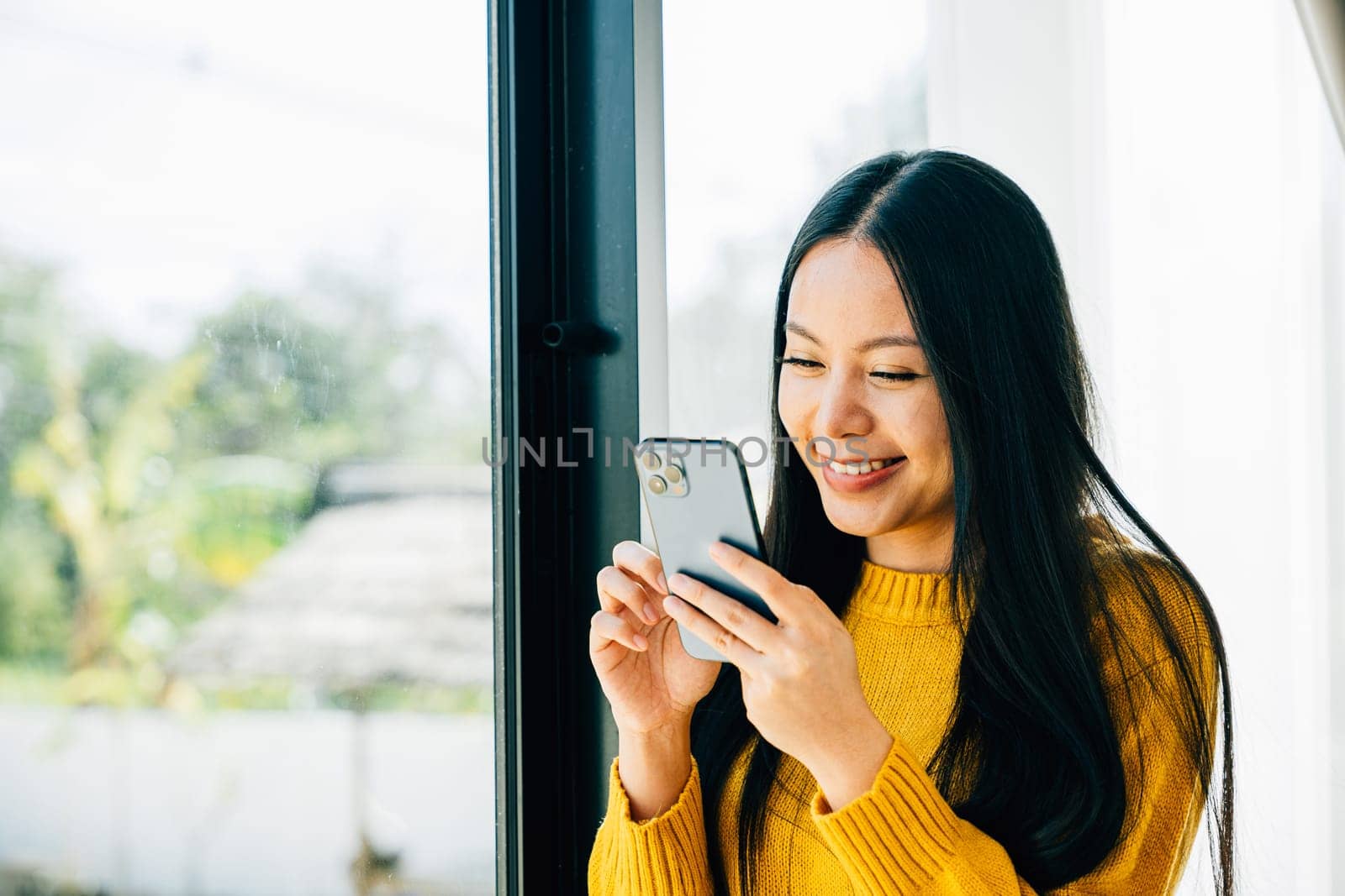 A woman with a smartphone app stands by the window typing and connecting for social media by Sorapop