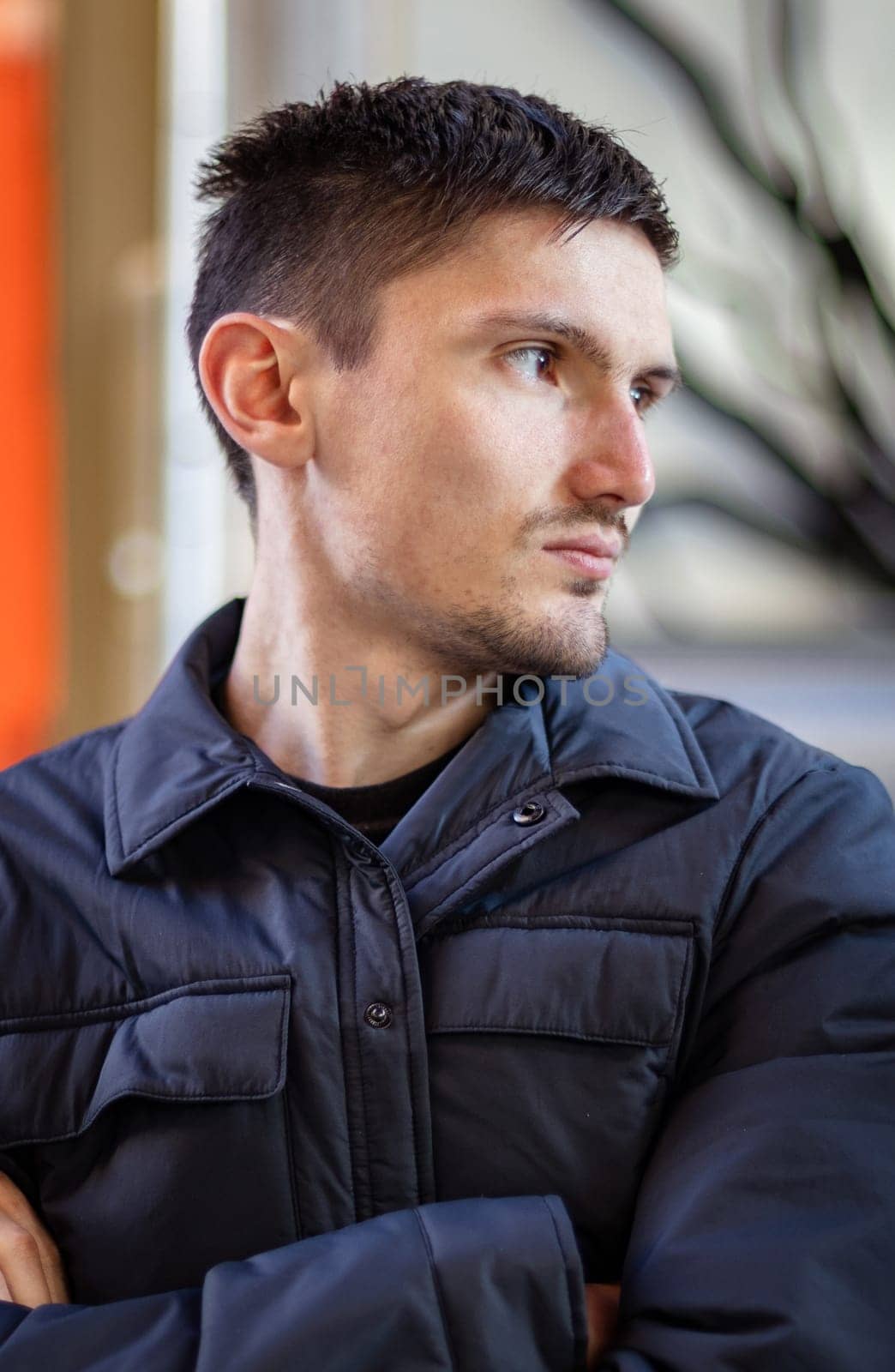 Portrait of one young handsome caucasian brunette guy in a dark fashionable jacket sitting at the table of the street terrace of the restaurant looking to the side, close-up side view.