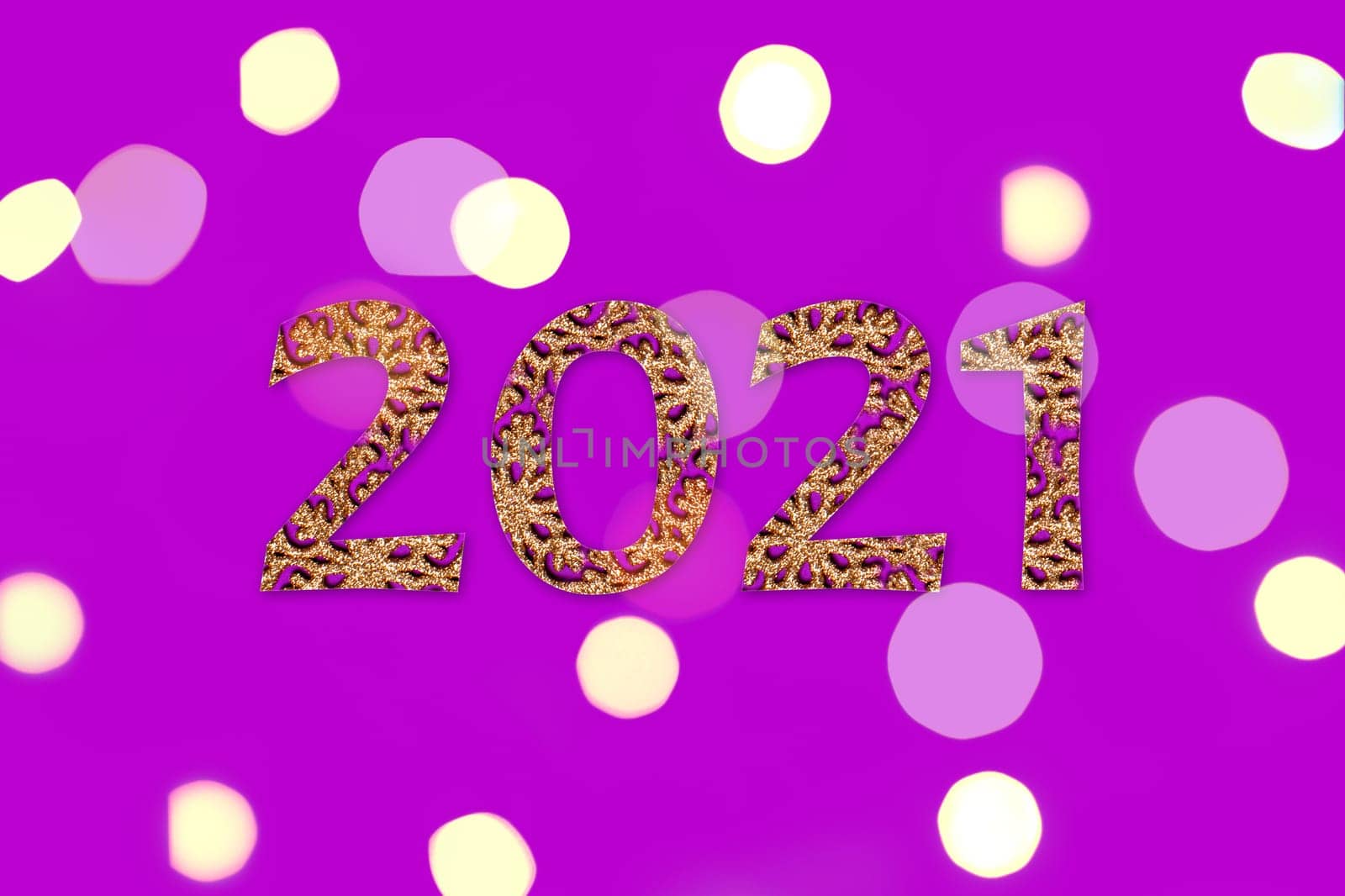 Christmas inscription 2021 laid out of gold Christmas decorations on a purple background. Surrounded by yellow lights.