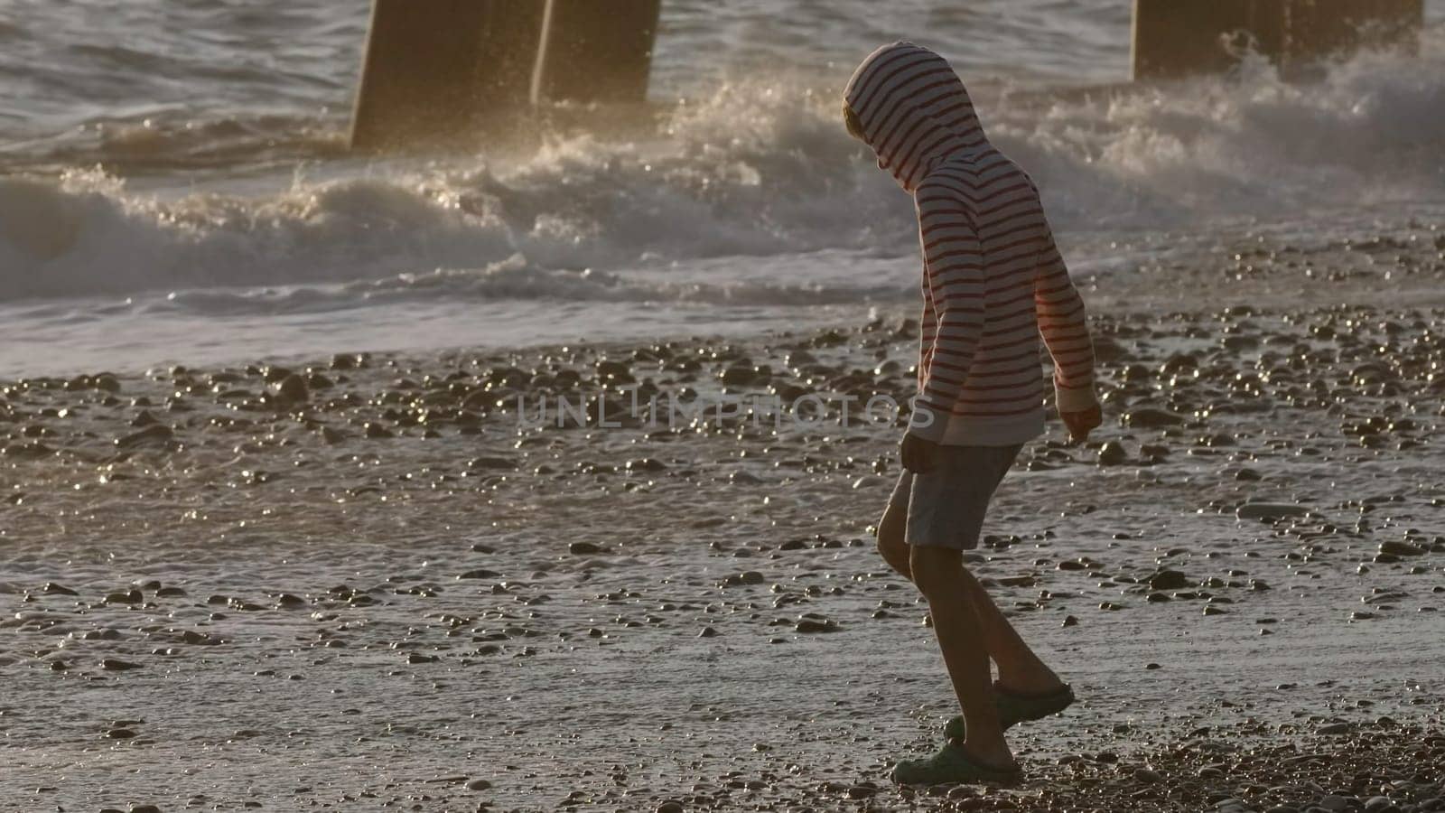 Boy walking by the sea, enjoying summer vibes. Creative. Boy child on a pebble beach in slow motion during sunset. by Mediawhalestock