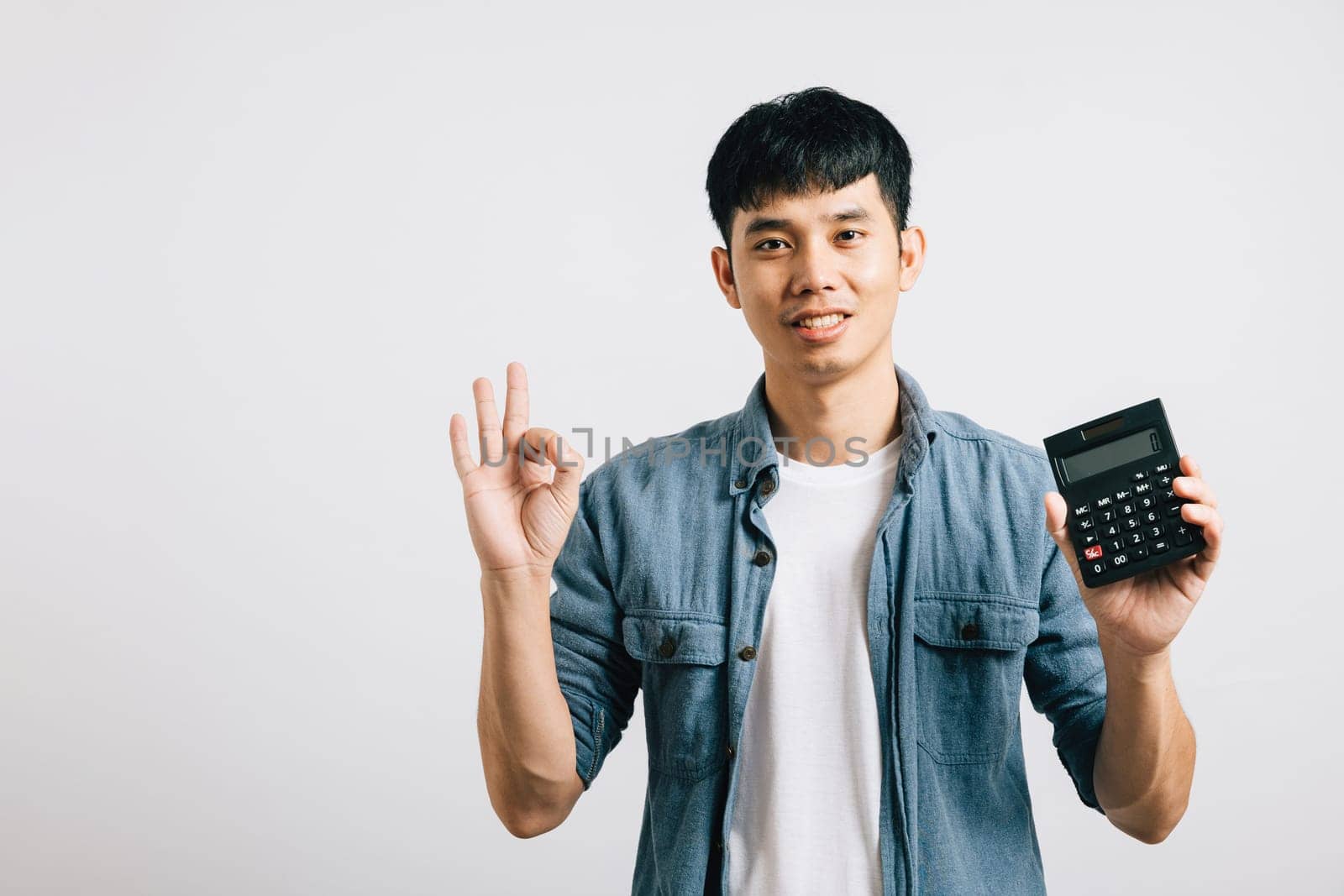 Portrait of a young man isolated on grey, showcasing a calculator, cash savings, and flashing OK sign finger. It's a TAX day concept, signifying financial success and responsible expense management.