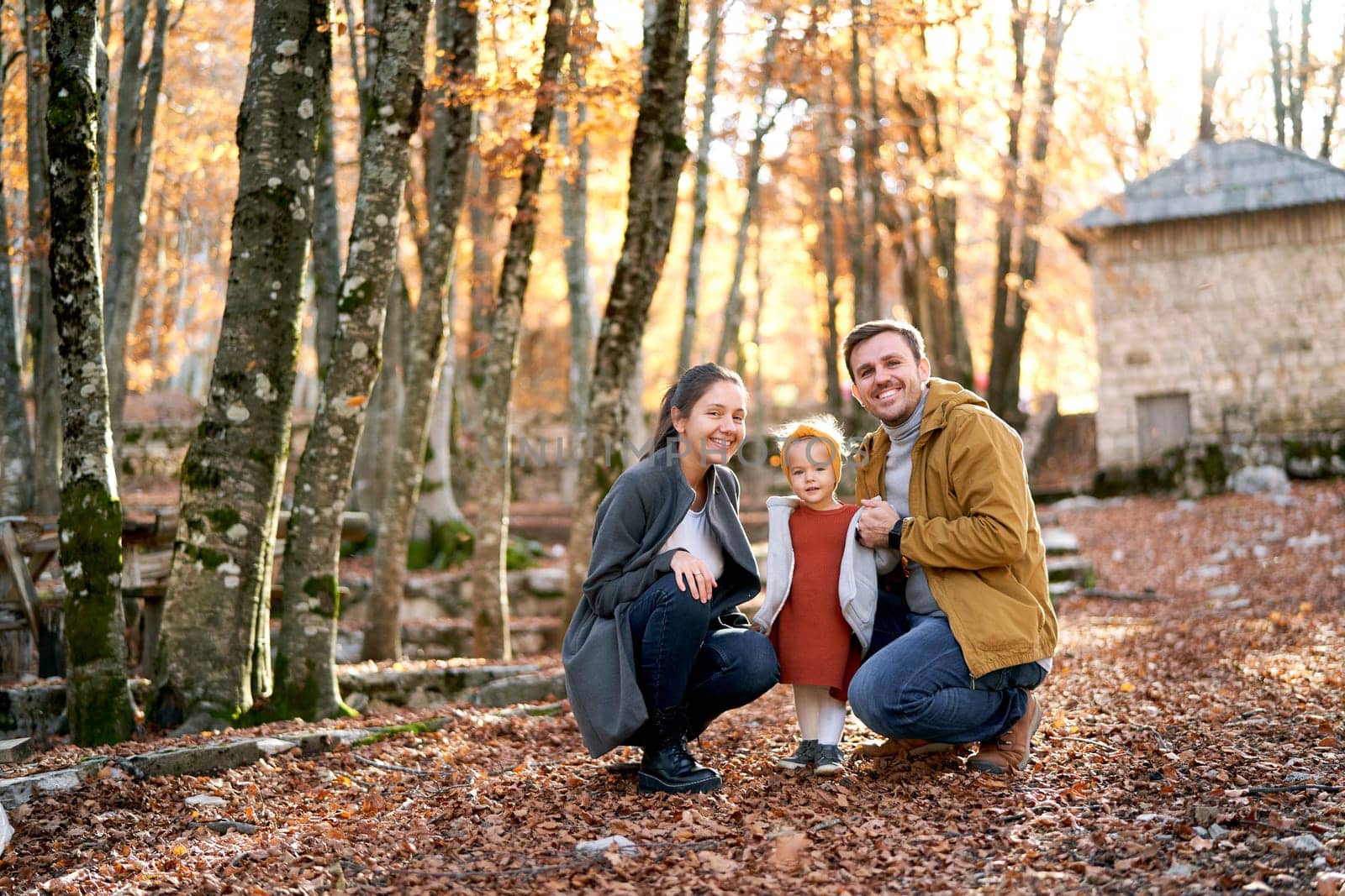 Smiling dad and mom are squatting near a little girl among fallen leaves in the forest. High quality photo