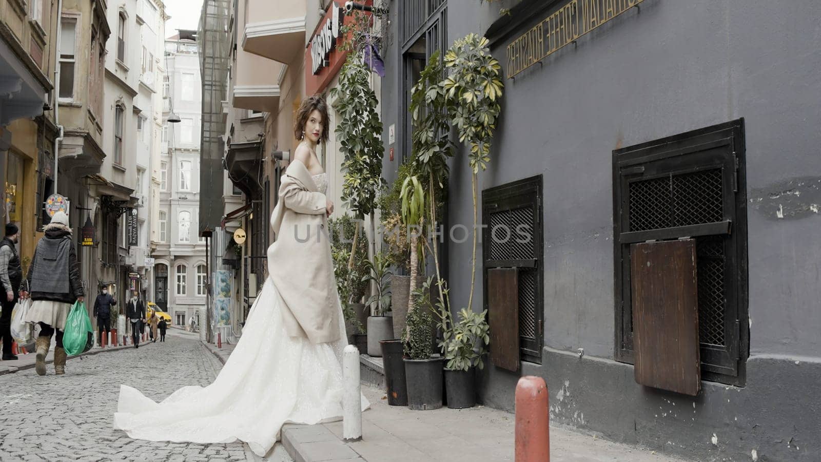 Turkey, Istanbul - July 27, 2022: Young fashionable luxury girl bride in elegant long wedding white dress in the narrow street of the old city. Action. Brunette woman standing during wedding photo shooting. by Mediawhalestock