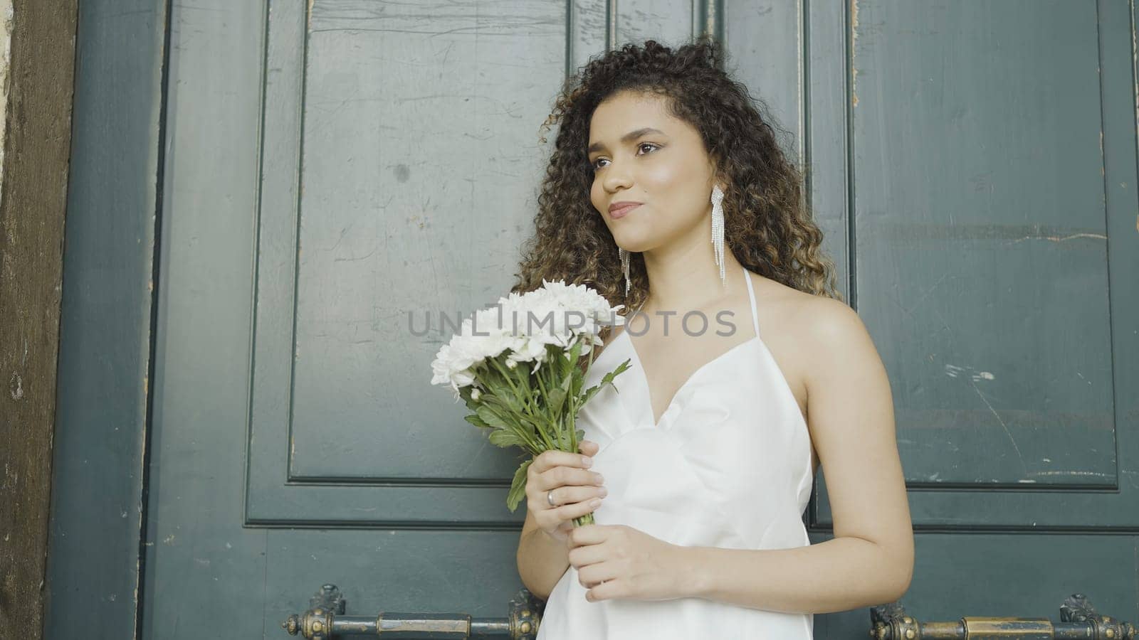 Beautiful young woman with bouquet of white flowers on street. Action. Attractive young woman smiles and holds bouquet of flowers. Smiling woman in dress and bouquet of flowers is waiting for date by Mediawhalestock