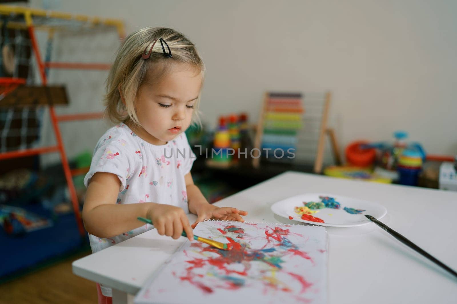 Little girl draws in an album with a brush with paints sitting at the table. High quality photo