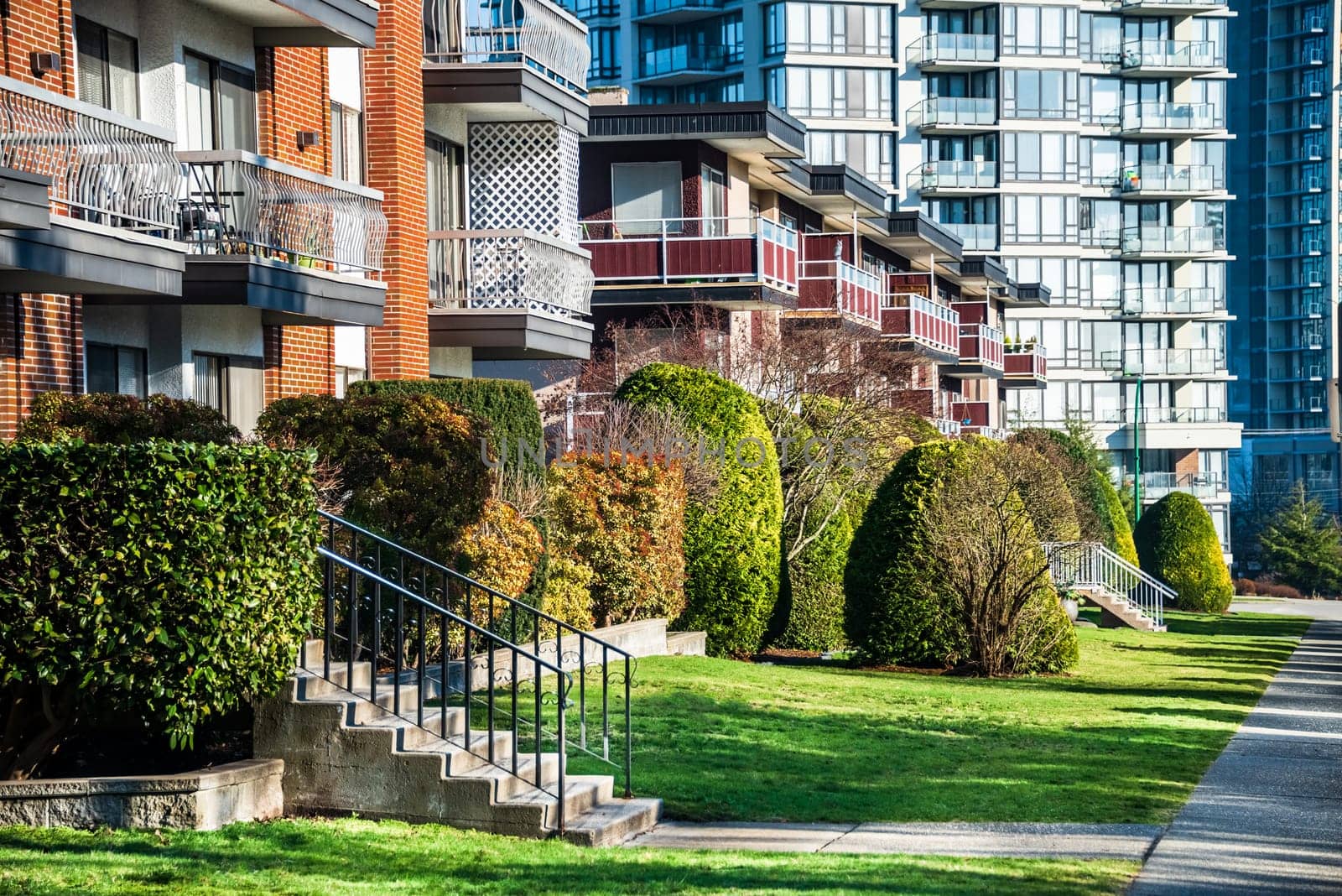 Facades of residential apartment buildings with doorsteps from the green lawn in front. Low-rise condo building on high-rise tower backgfound