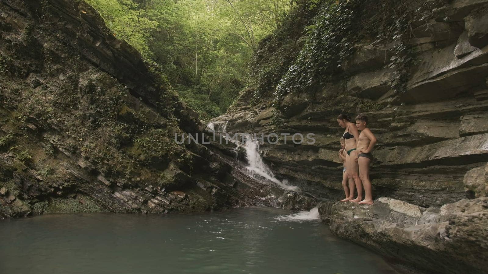 Woman with children by pond in forest. Creative. Natural waterfall with pond and bathing tourists. Family with children swim in natural pond with waterfall in forest.