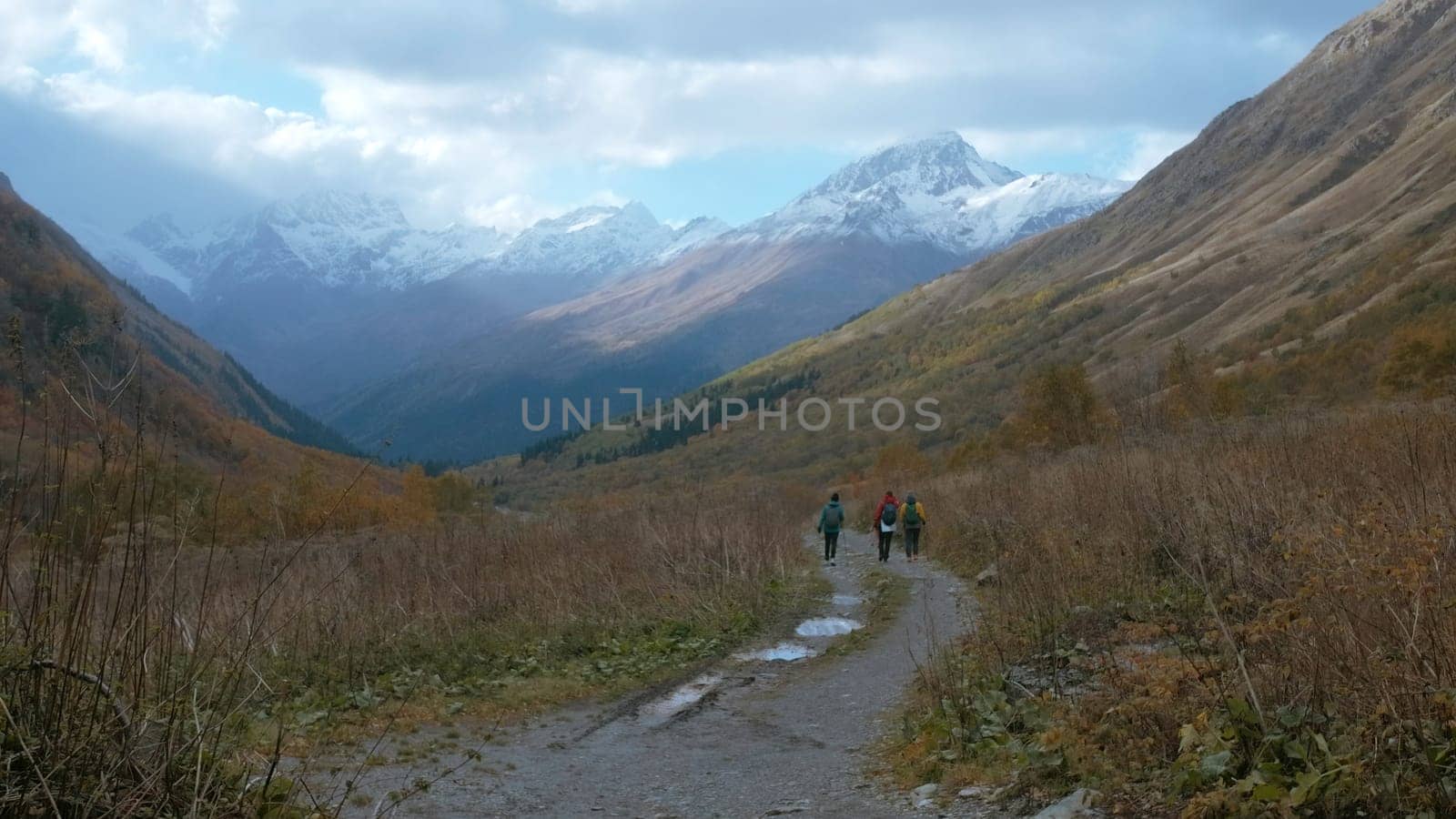 Tourists walk along trail in mountain valley in autumn. Creative. Hiking in mountain valley on cloudy autumn day. Trail in mountain valley with beautiful landscape of snowy peaks and cloudy sky.