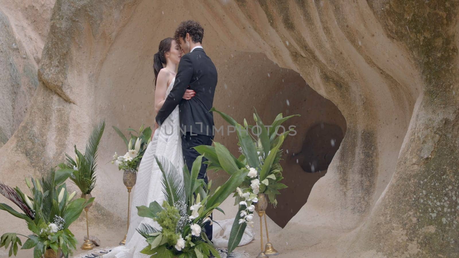 Beautiful newlyweds on rock background. Action. Hugging newlyweds on background of cave during snow. Wedding ceremony at rocks during snowfall by Mediawhalestock