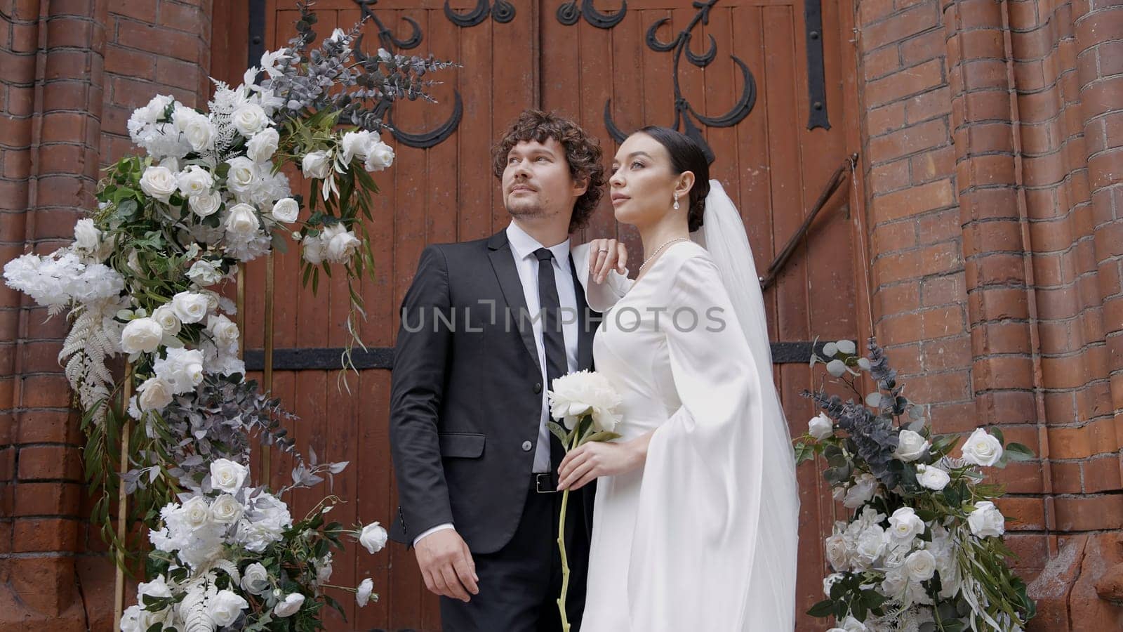 A bright and spectacular wedding couple. Action.Newlyweds where a bride in a white dress with a slit on her leg and a man with a beard in a suit walking next to a white arch with fresh flowers. High quality 4k footage